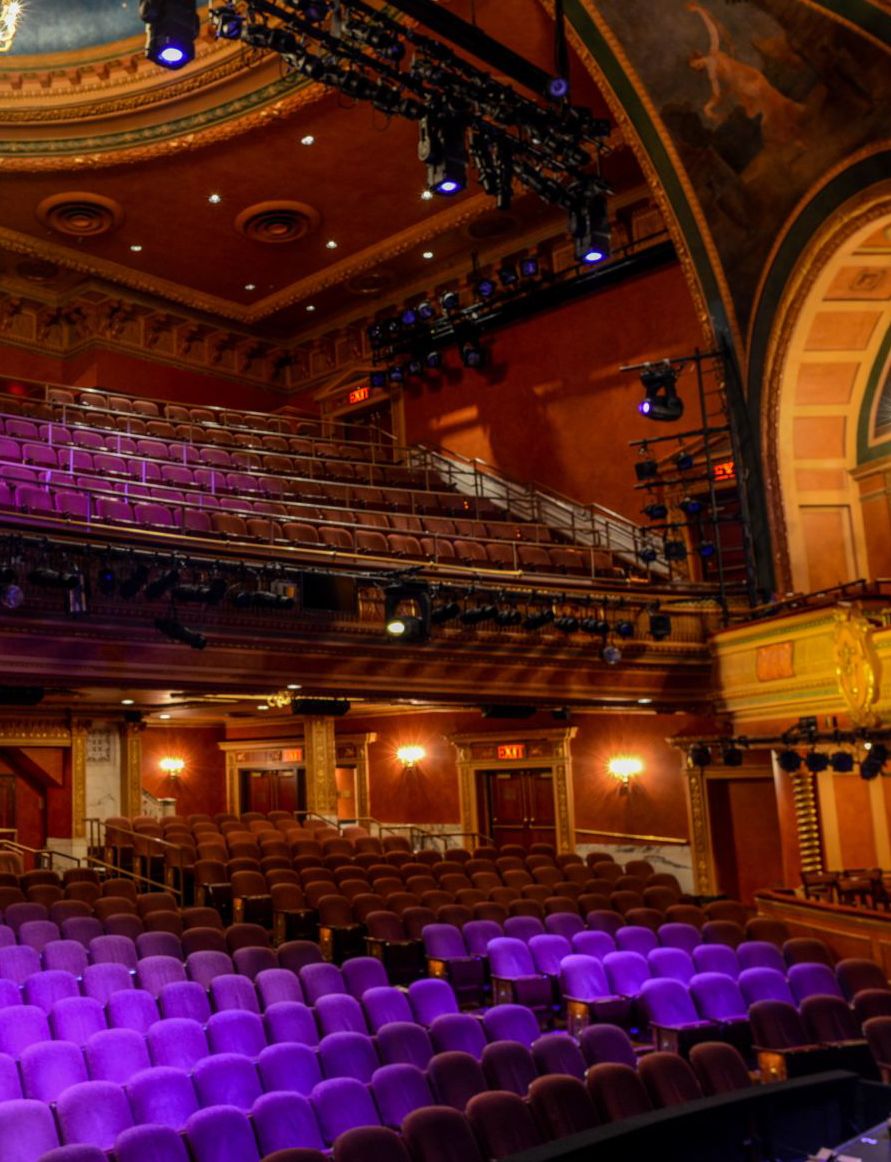 interior of the American Airlines Theatre taken from the edge of the stage and facing the seats. The seats are purple and the tone of the wood is yellow. Both the mezzanine and orchestra are shown.