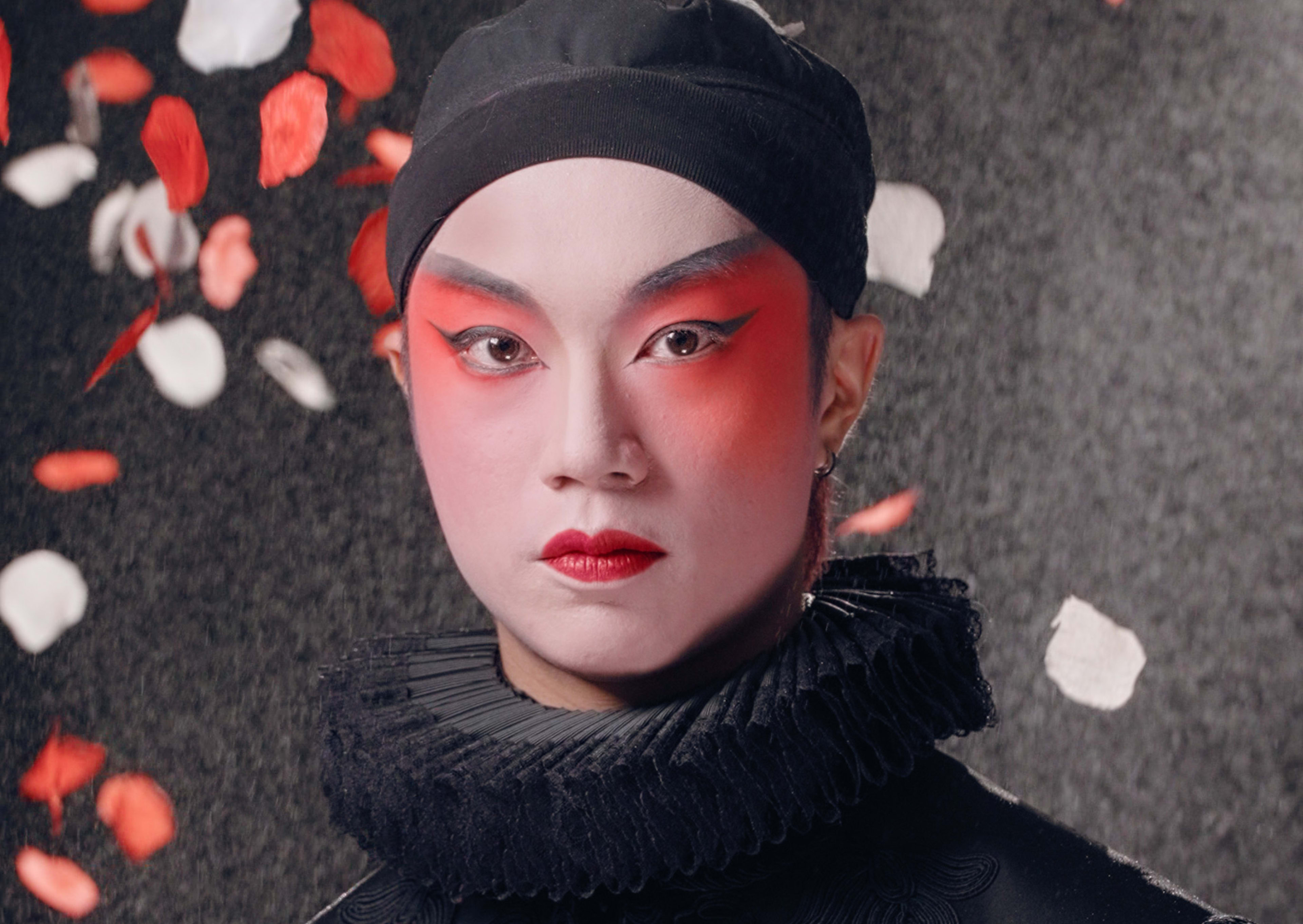 天台製作當代劇場作品《曲解莎劇》（History Play）主視覺海報。演員梁俊仁飾演 Rizzly（Southampton伯爵），他面容沉靜，化著大膽且具前衛感的舞台妝容，深紅色眼影與紅唇對比強烈。他身穿精緻的黑色伊莉莎白風格服裝，頸部佩戴黑色拉夫領（ruff collar）。背景為充滿質感的深色調，紅、白玫瑰花瓣在四周飄落，營造出強烈的戲劇氛圍與香港當代劇場的獨特美學。《曲解莎劇》讀劇演出。