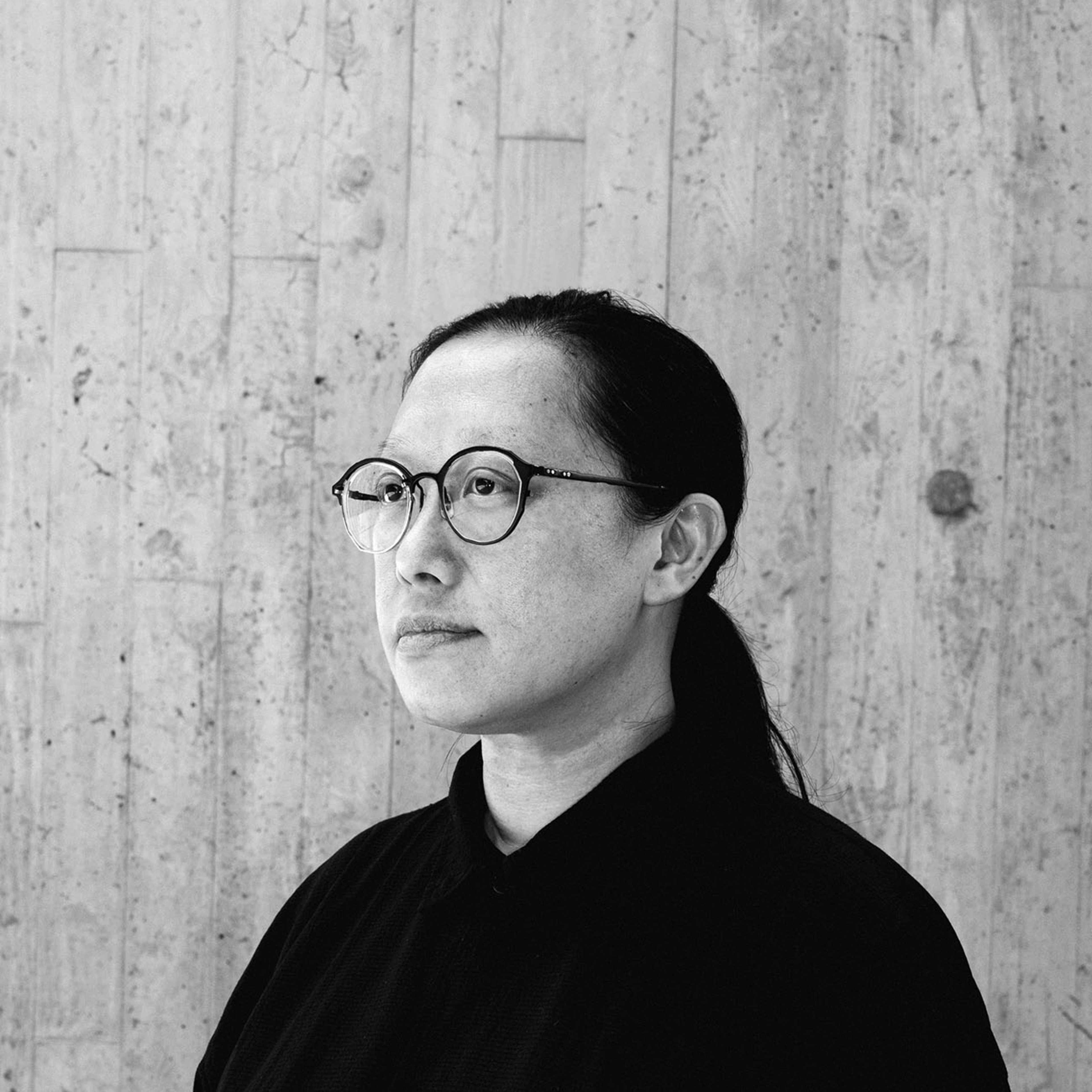 A contemplative black-and-white headshot of performer Bobo Lee (李筱怡), captured against a raw, textured concrete wall characteristic of Hong Kong's contemporary black box theatre aesthetic. Bobo Lee is shown in profile, wearing dark-rimmed glasses and a black shirt, embodying the minimalist and avant-garde spirit of Rooftop Productions (天台製作). A headshot.