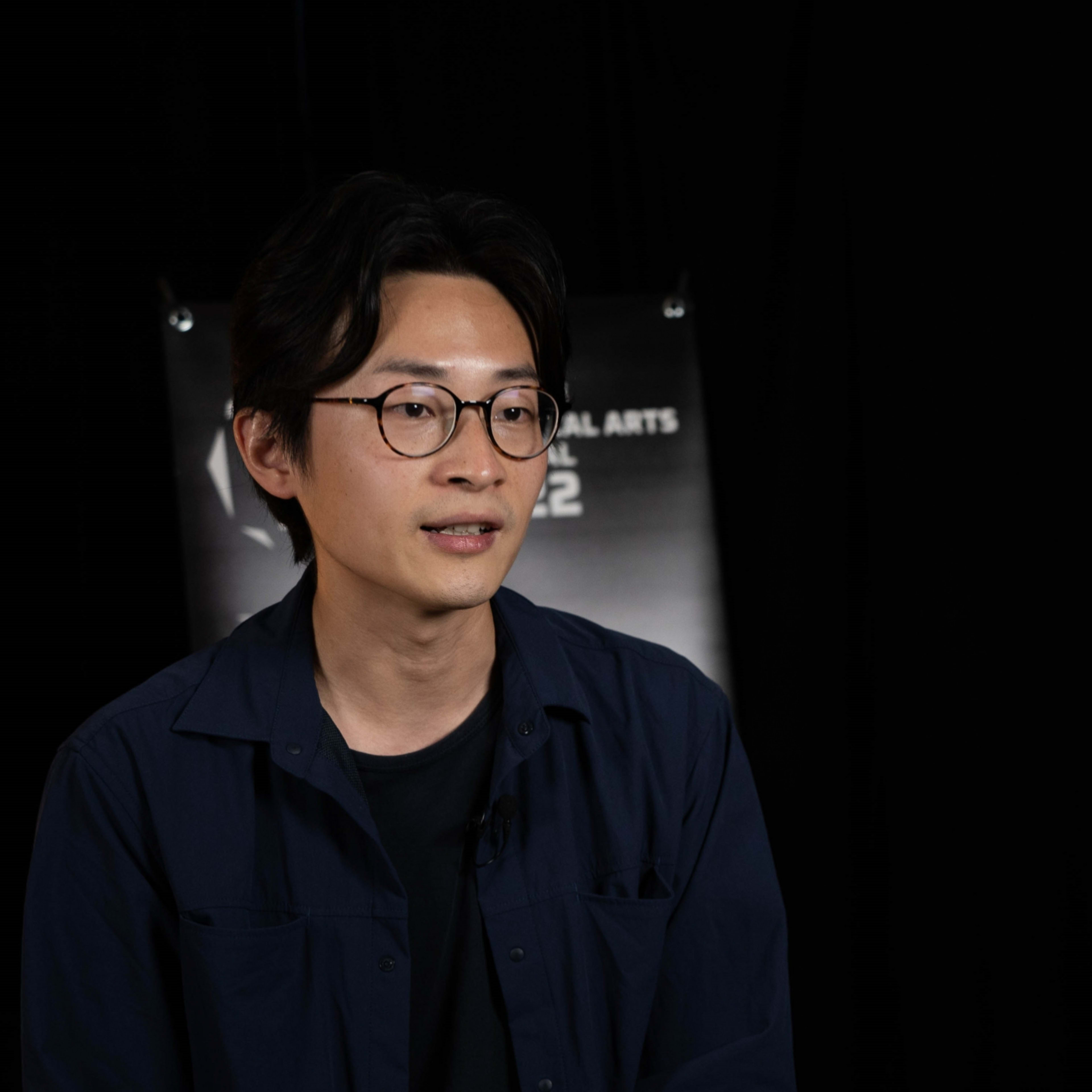A studio headshot of Hong Kong theatre actor Fong Hiu-tan, who is wearing round-rimmed glasses and a dark blue button-down shirt. He is captured in a soft, focused light against a dark background, reflecting the professional and artistic atmosphere typical of Rooftop Productions (天台製作) theatre work. A headshot.
