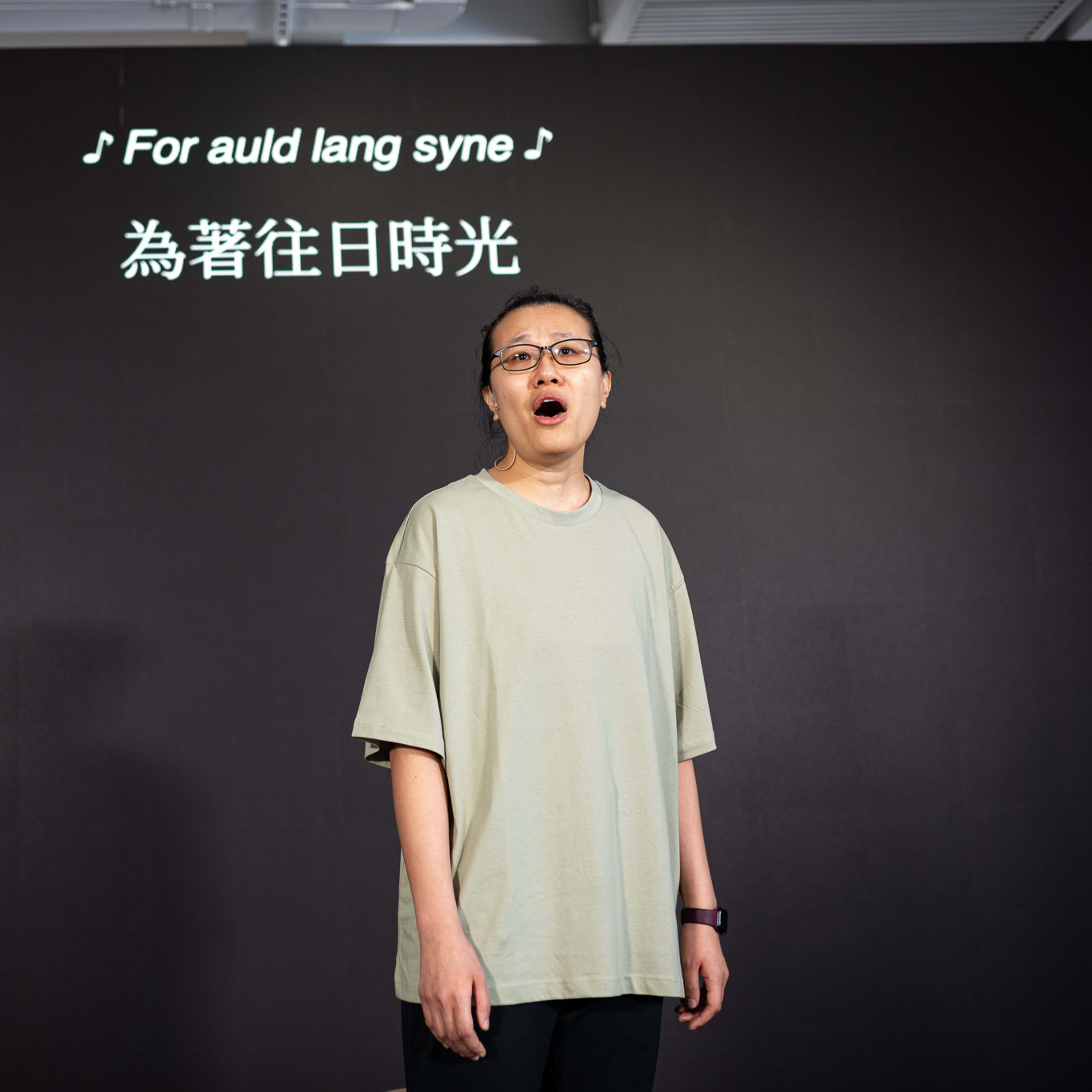 In a rehearsal for the contemporary Hong Kong theatre production 'Karaoke' (《卡啦OK》) by Rooftop Productions (天台製作), performer Michelle Li stands on a minimalist stage against a stark black backdrop. She is wearing a simple light-colored t-shirt and glasses, singing with visible emotion. To her left, a bright blue costume hangs on a rack. Projection text above her head reads: "For auld lang syne". As she sings, she captures the poignant essence of the performance: "For auld lang syne."