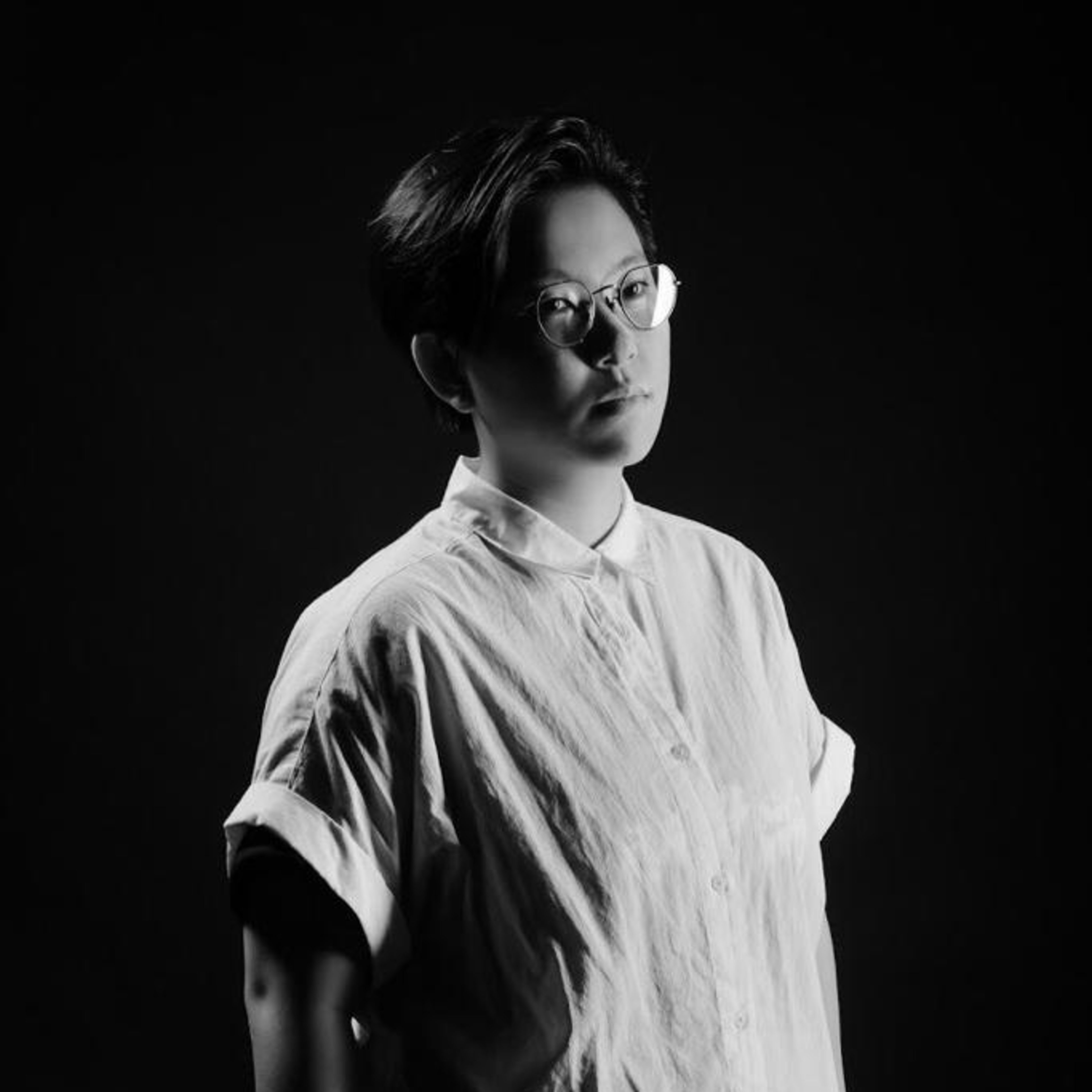 A dramatic black-and-white headshot of performer Lai Ka-ki for a Rooftop Productions (天台製作) theatrical performance. She wears glasses and a short-sleeved, collared shirt, captured against a stark dark background with high-contrast studio lighting that emphasizes a contemplative, focused expression. A headshot.