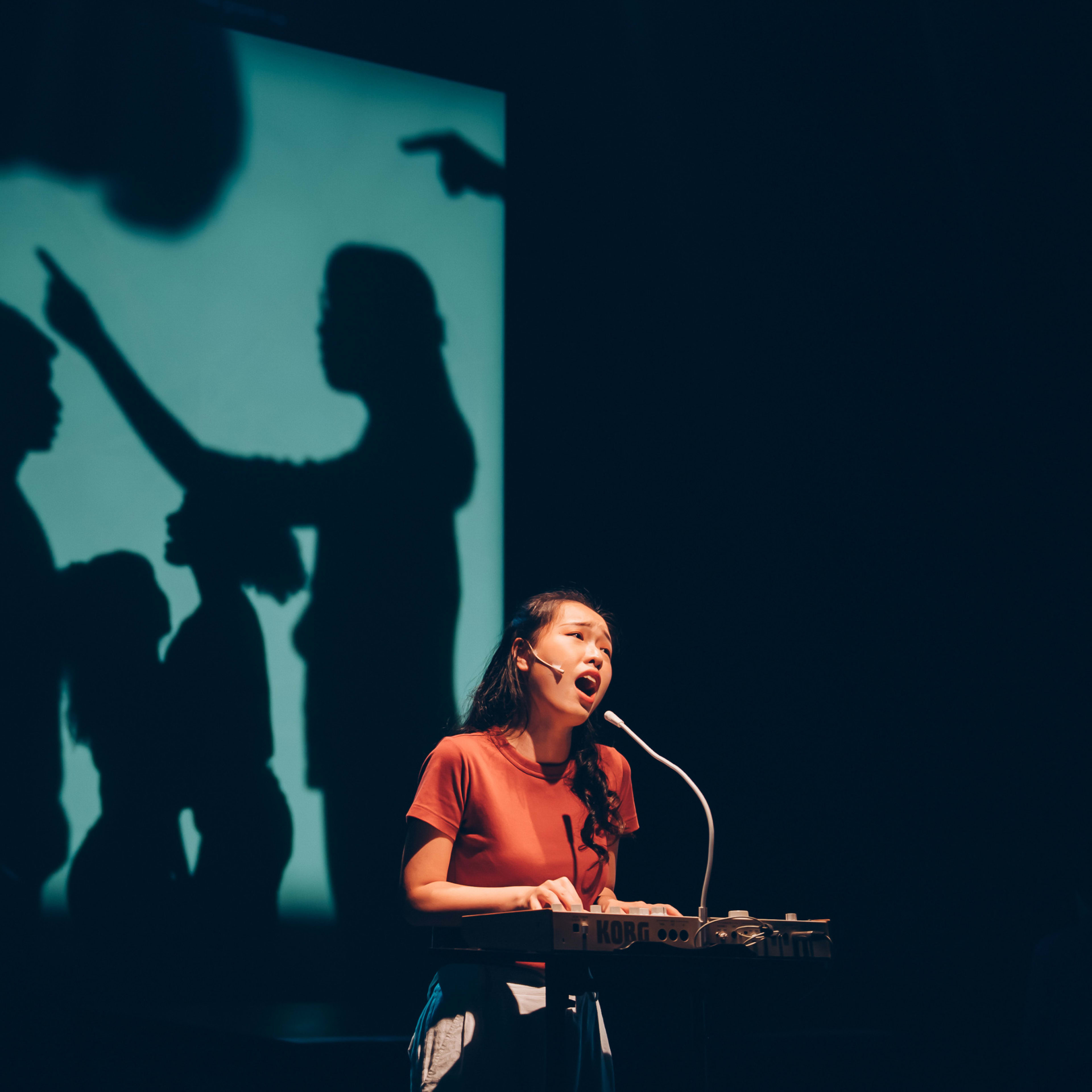 Sum Cheuk-yiu, performing live in the contemporary Hong Kong theatre production 'Songs of Innocence and Experience', sings expressively into a microphone while playing an electronic keyboard. She is brightly lit on a dark stage, against a large teal-blue screen projecting dramatic silhouettes of figures interacting, one pointing an accusatory finger. In the shadowy background, a musician plays an electric guitar, adding to the multimedia stage performance. 'Your daughter has grown up.'
