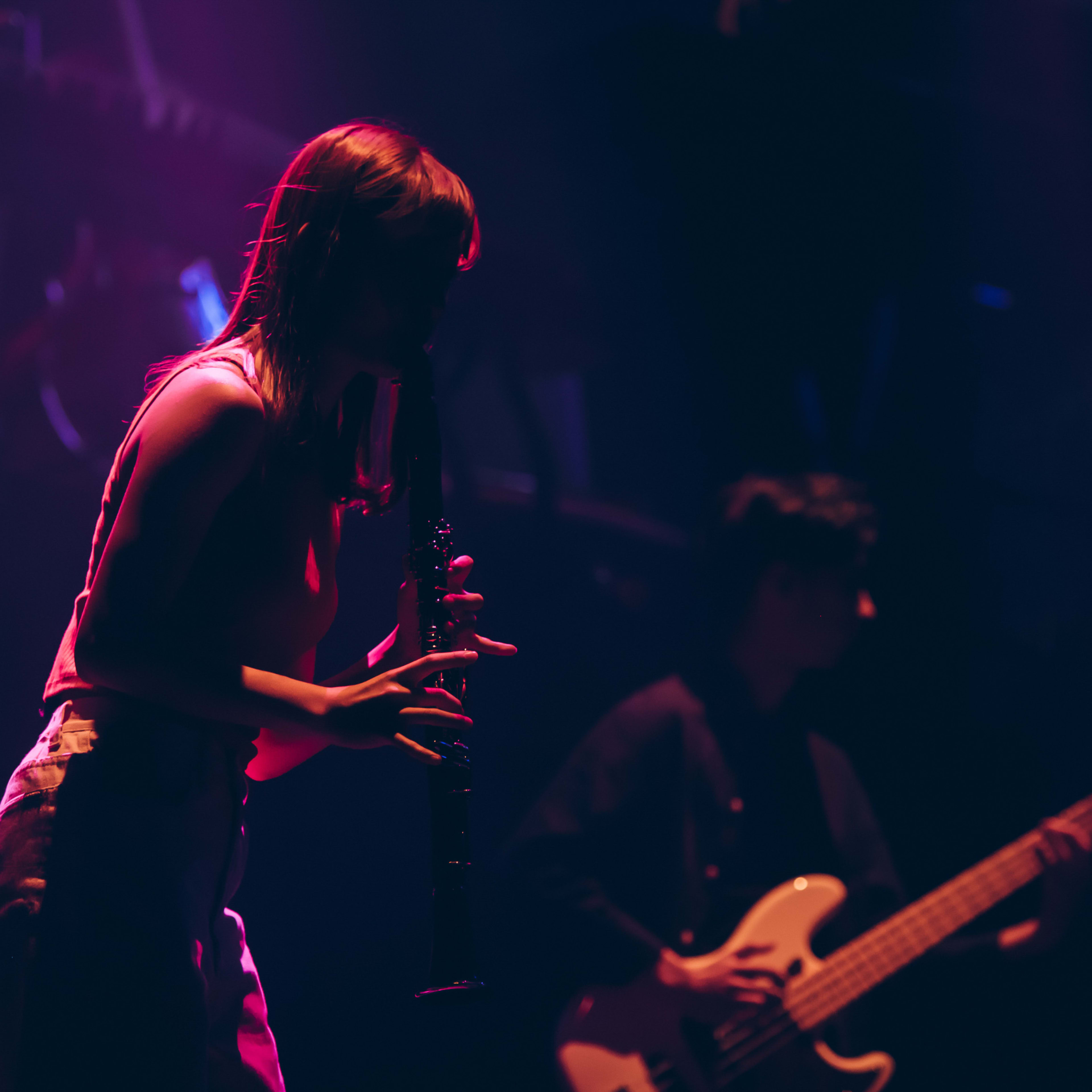 In a moody, red-lit stage production of Rooftop Productions' contemporary theatre piece 'Songs of Innocence and Experience', Sin Lok-yan stands in silhouette playing the flute, while band leader Wu Lui-fung plays bass guitar in the background. This atmospheric performance captures the intensity of Hong Kong live stage art. As Sin Lok-yan delivers a poignant moment of the show, she sings, "No money no use, no money no dream."