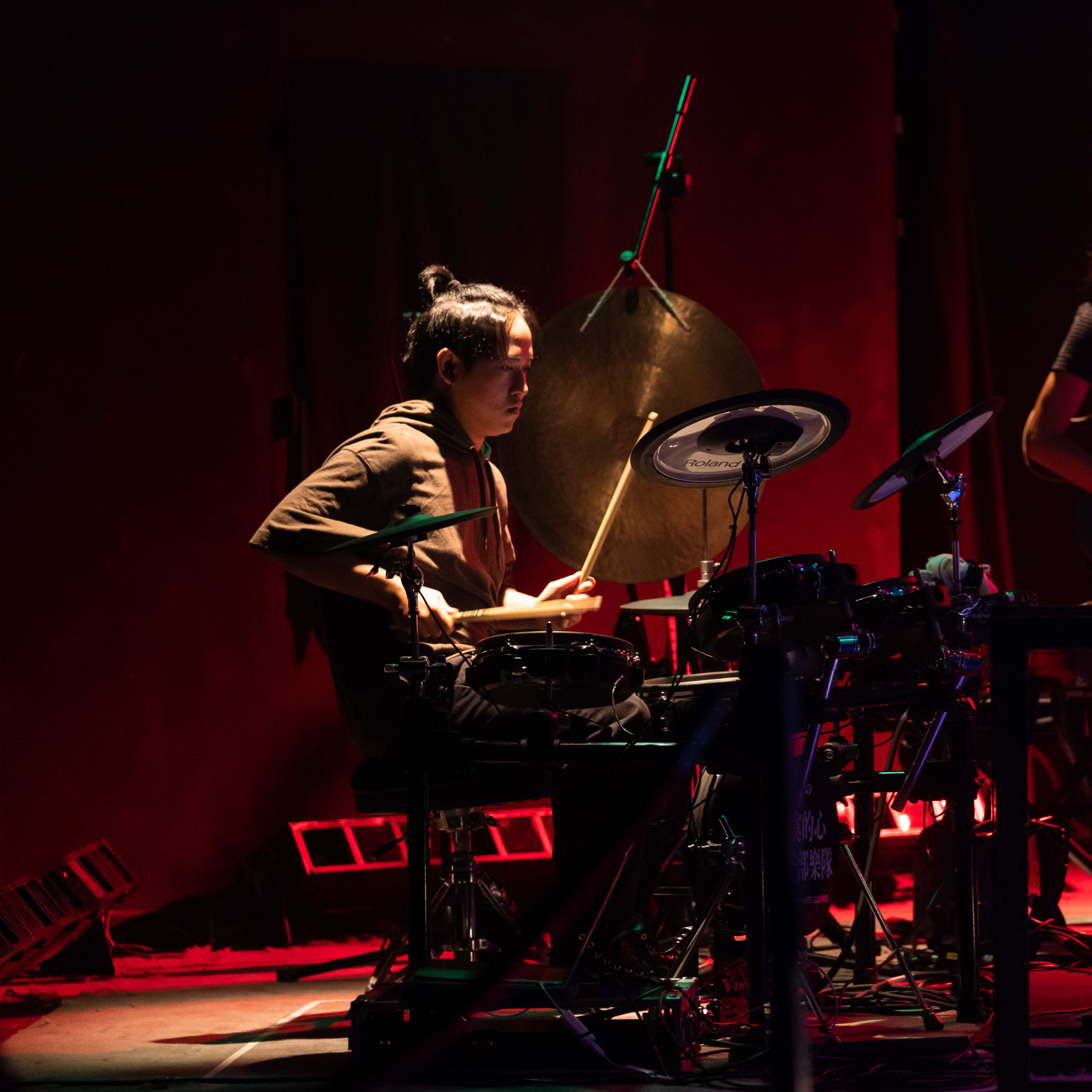 In a dramatic scene from Rooftop Productions' contemporary theatre performance, 'Songs of Innocence and Experience,' Sung Kwok-tung is seen focused on playing electronic drums and a large gong under moody, deep red stage lighting, while Chin Wing-kar plays an electric guitar in the background. This atmospheric production explores complex themes of musical storytelling in Hong Kong theatre. As the tension mounts, a performer sings, "Don't cry out loud."