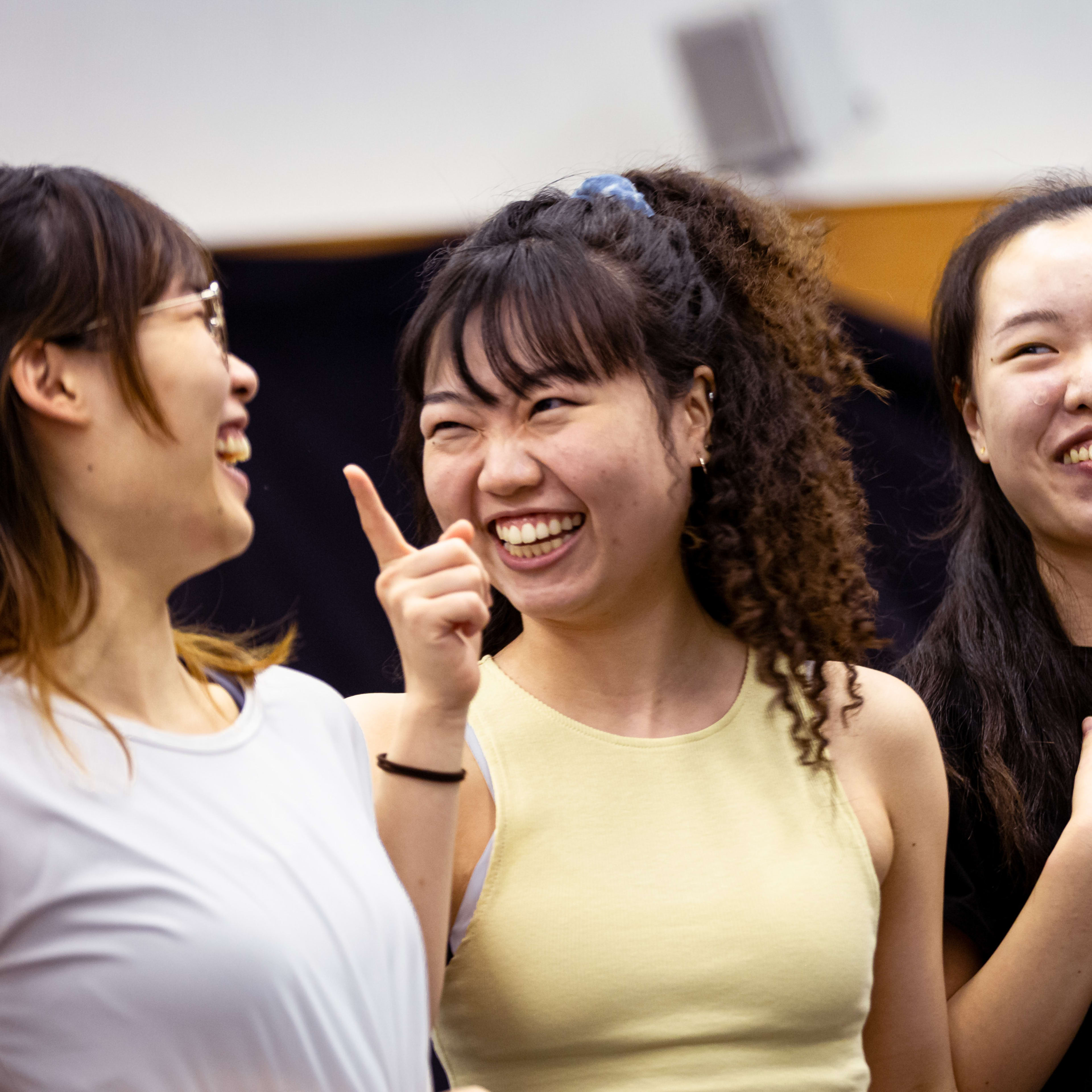 Three performers—Lee On-sang, Sum Cheuk-yiu, and Lau Ka-ying—share a lighthearted moment during a rehearsal for the Rooftop Productions contemporary theatre piece, 'Songs of Innocence and Experience'. Sum Cheuk-yiu (playing Abdiel) points playfully while laughing, flanked by Lee On-sang (playing Moloch) and Lau Ka-ying (playing Belial), who expresses heartfelt joy with her hands over her chest. The scene captures the collaborative spirit of HK stage performance. Songs of Innocence and Experience Rehearsal.