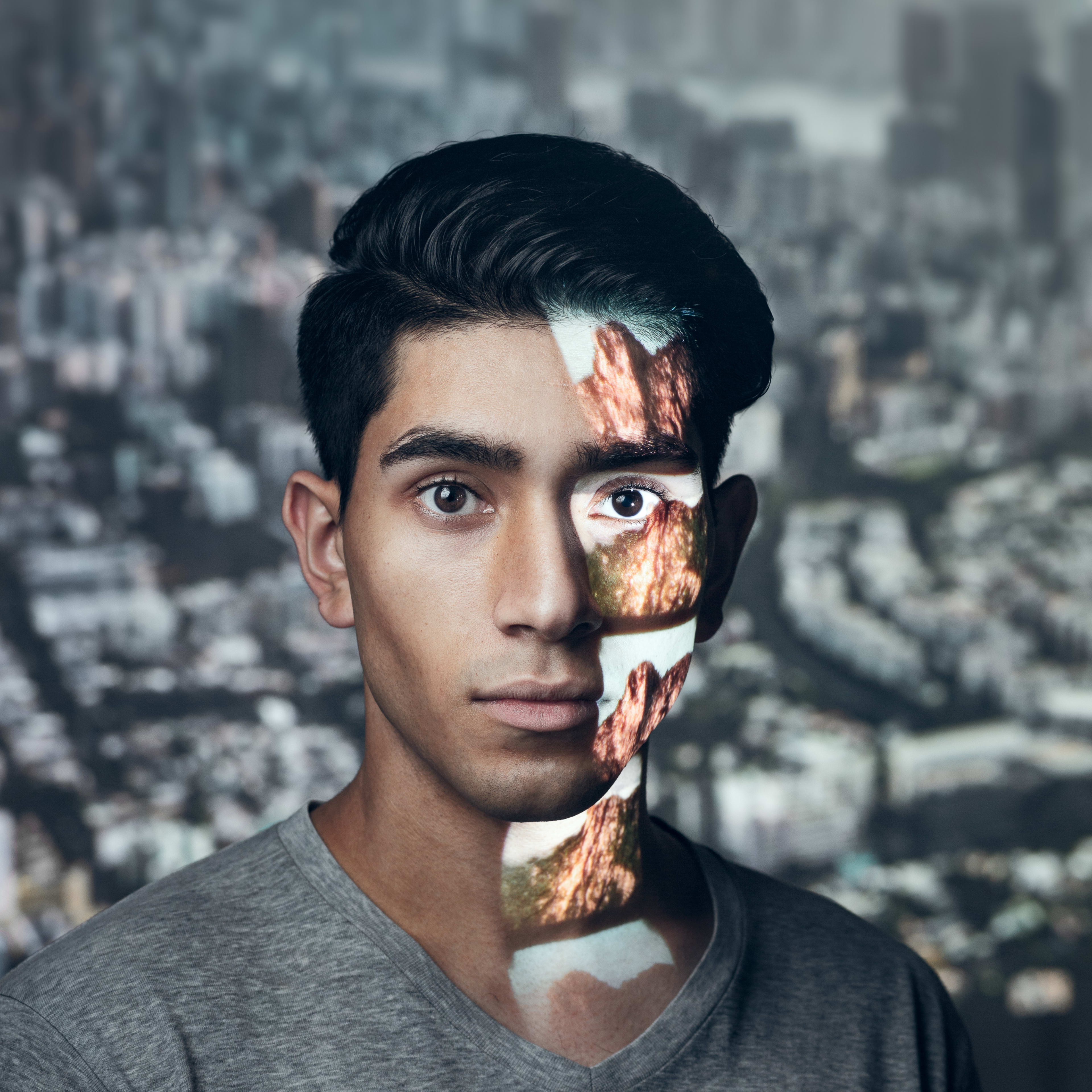 天台製作（Rooftop Productions）當代劇場作品《山下的證詞》宣傳劇照。演員巫加沙面部右側投射著山巒的影像，與背景中模糊的城市景觀形成對比，展現出香港當代劇場獨特的敘事風格與深刻的意象。演員表情沉靜，體現了劇作發人深省的氛圍。《山下的證詞》