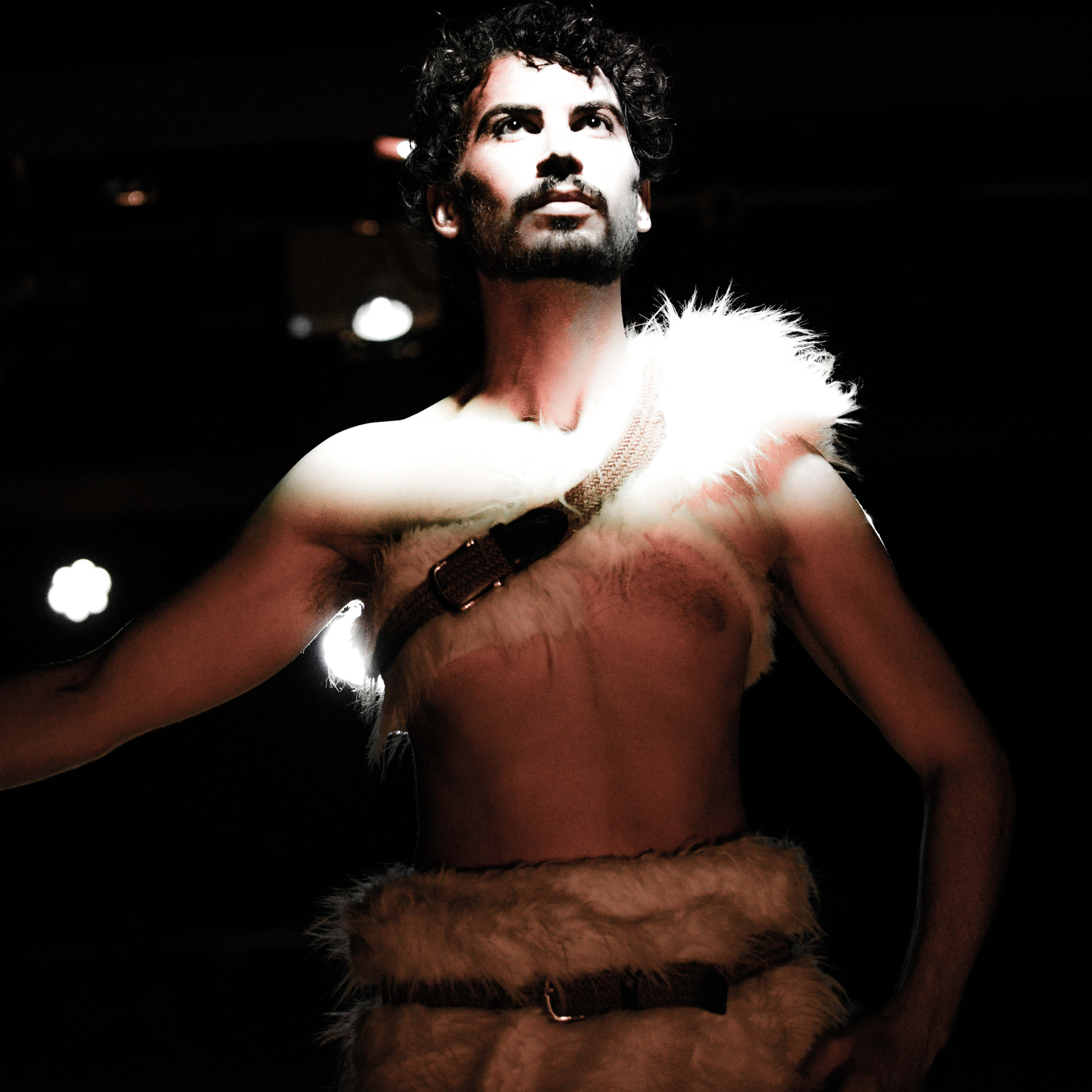 A dramatic stage shot from Rooftop Productions' contemporary theatre performance, The Beautiful Ones. Actor Ivor Houlker stands center stage, captured in a dynamic pose. He is shirtless, dressed in a primitive-style faux fur wrap secured by a belt, illuminated by sharp, high-contrast spotlighting against a dark, moody background. His expression is intense and focused, embodying a raw, primal energy characteristic of Hong Kong's experimental stage. 'The Beautiful Ones'