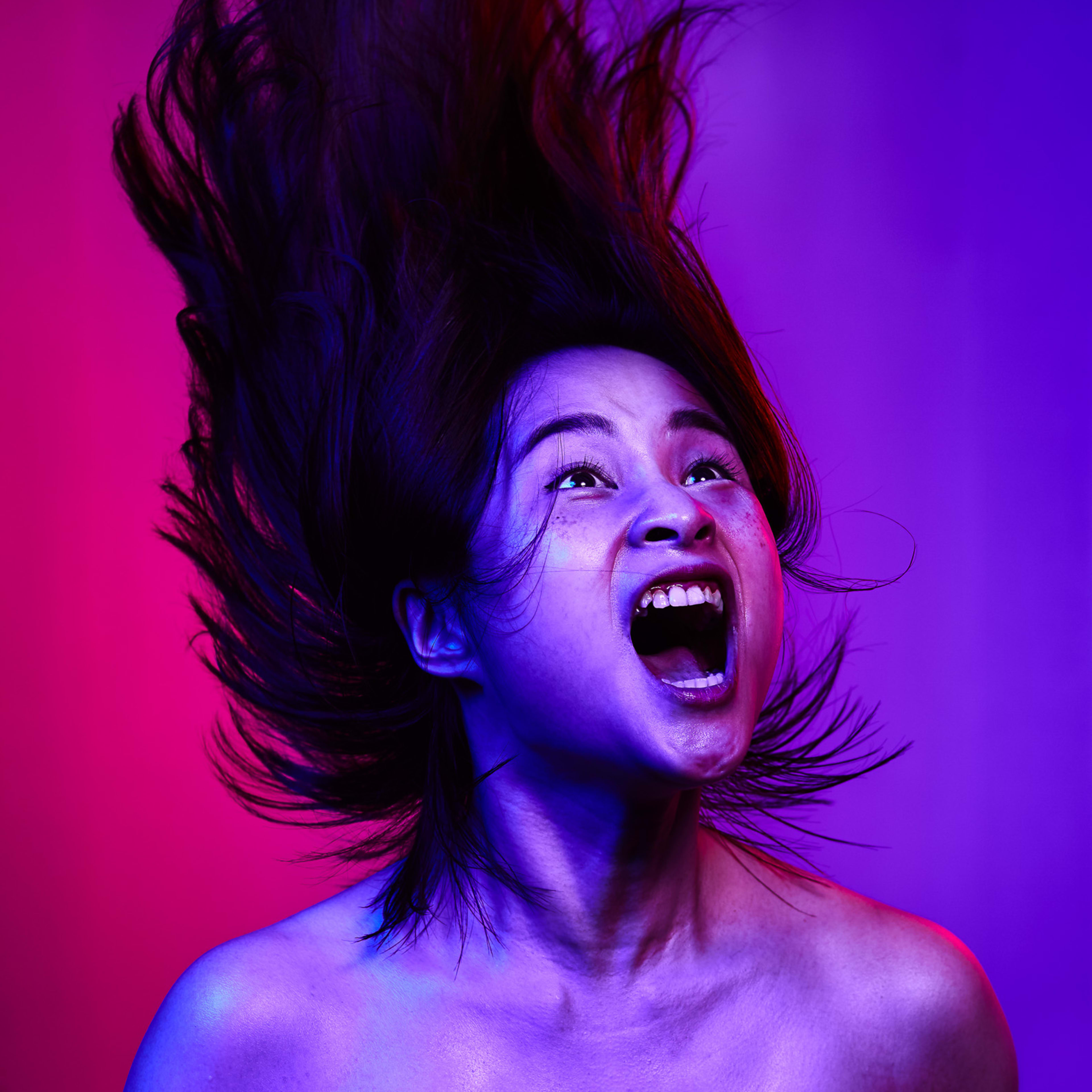 A dramatic promotional portrait of actor Lung Jes for the Rooftop Productions contemporary theatre piece, The Furies Variations. Captured against a vibrant dual-tone backdrop of magenta and electric blue, the subject is mid-motion, hair thrashing upward while emitting a raw, intense scream that embodies the visceral energy of the performance. Promotional photo of Lung Jes for The Furies Variations.