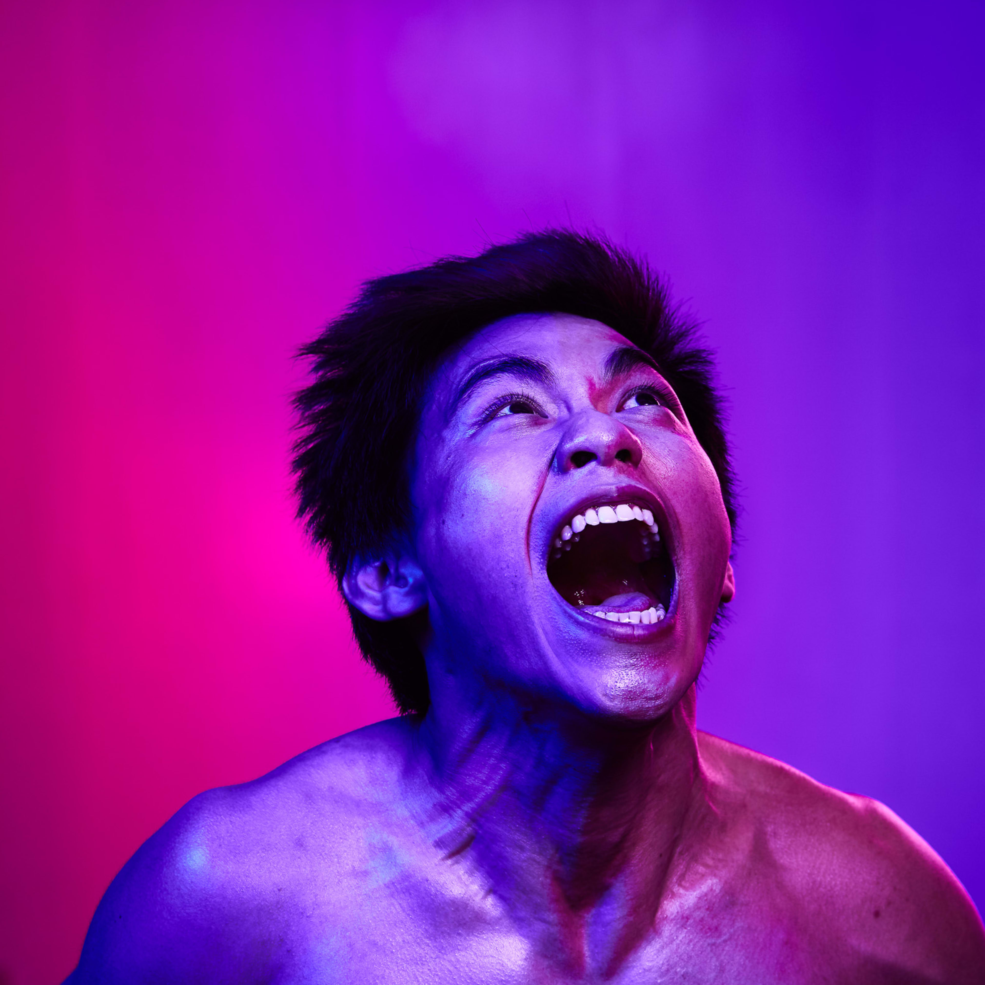 A dramatic promotional portrait of actor Yau Fuk Wing for Rooftop Productions' contemporary theatre piece, The Furies Variations. Yau is captured from the chest up, head tilted back with his mouth wide open in a raw, primal expression of intense emotion. The scene is bathed in striking dual-tone neon lighting, with vibrant magenta illuminating one side of his face and a deep, moody purple casting shadows on the other, reflecting the experimental and visceral nature of Hong Kong stage performance. Promotional photo of Yau Fuk Wing for The Furies Variations.