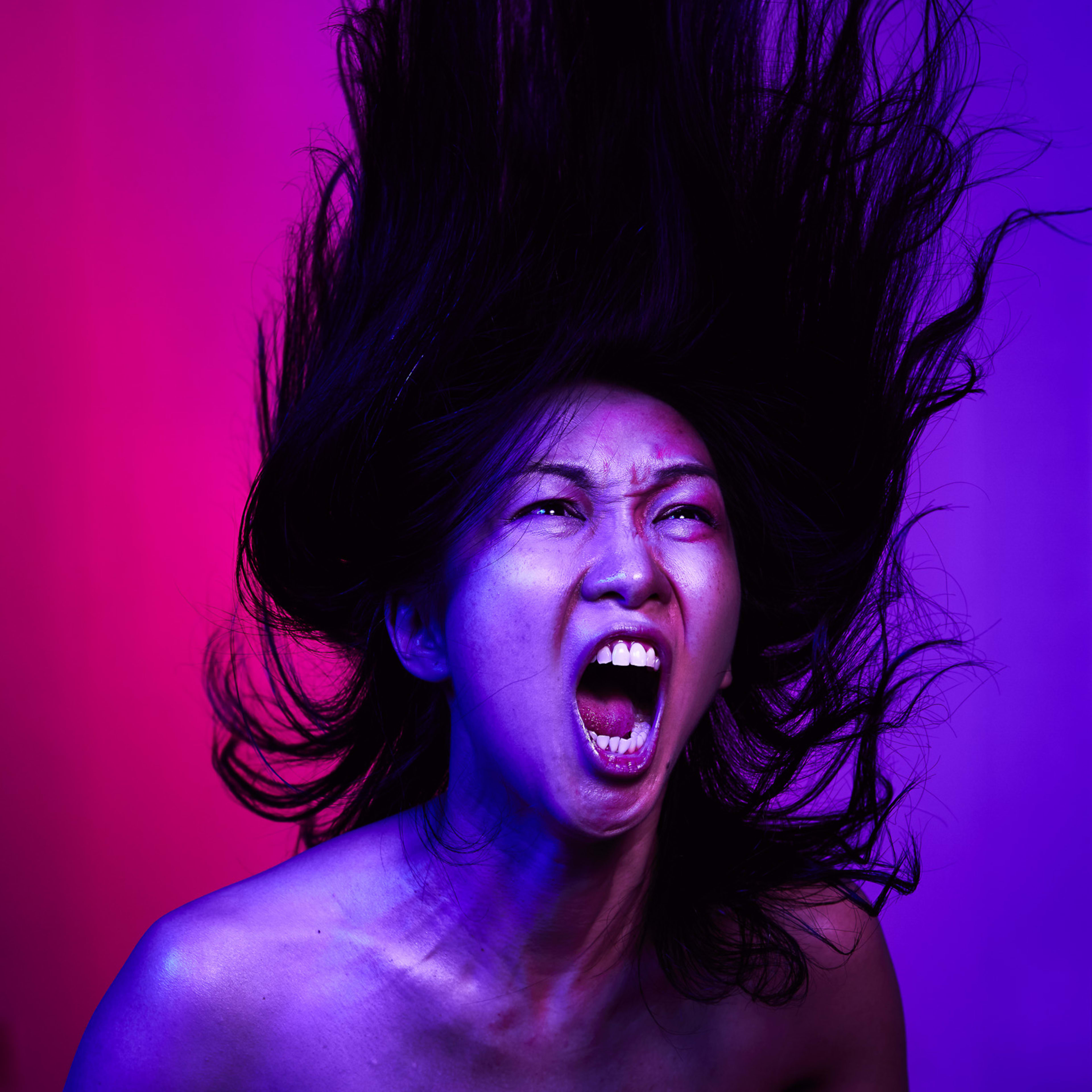 Promotional portrait of actor Maggie Chu for the contemporary Hong Kong theatre production, The Furies Variations. Captured against a striking dual-tone background of pink and purple, Chu is mid-action with her dark hair floating upwards in a dynamic, ethereal display. She expresses raw, visceral emotion, her mouth wide open in a fierce, powerful scream that embodies the intensity of the show. Promotional photo of Maggie Chu for The Furies Variations.