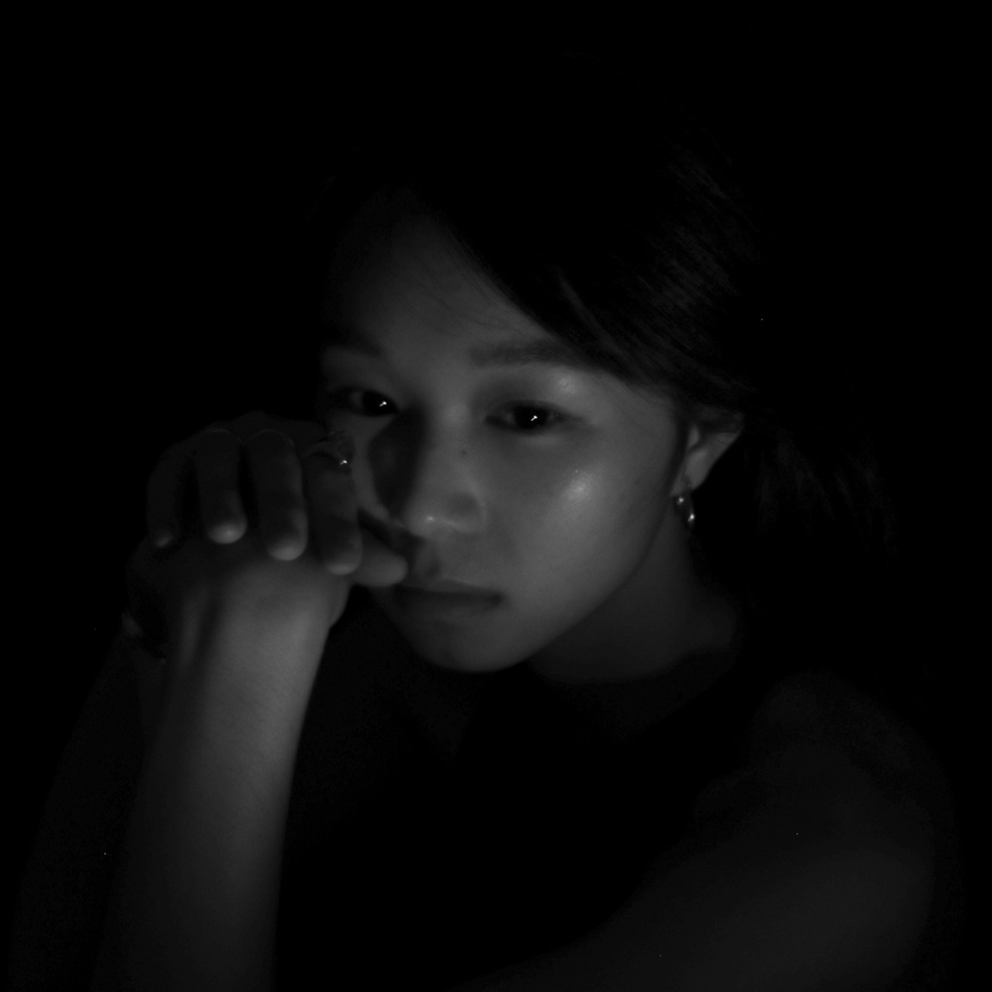 A dramatic close-up headshot of Hong Kong theatre performer Tiffany Lau, illuminated by low-key, moody lighting against a deep black background. She gazes intently toward the viewer, her hands held thoughtfully near her face in a gesture of quiet contemplation, capturing the raw, intimate aesthetic of a Rooftop Productions contemporary stage performance. A headshot.