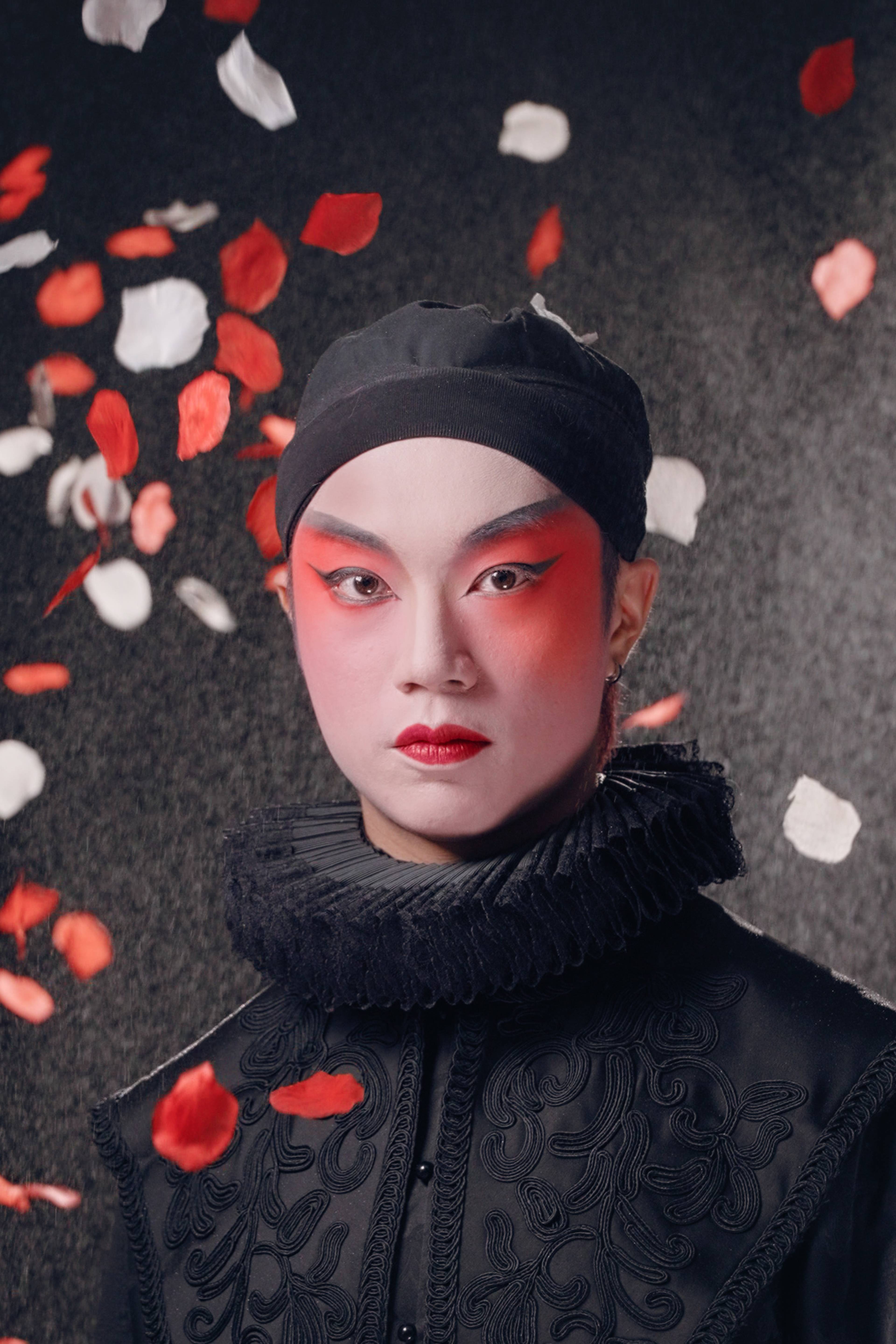 天台製作當代劇場作品《曲解莎劇》（History Play）主視覺海報。演員梁俊仁飾演 Rizzly（Southampton伯爵），他面容沉靜，化著大膽且具前衛感的舞台妝容，深紅色眼影與紅唇對比強烈。他身穿精緻的黑色伊莉莎白風格服裝，頸部佩戴黑色拉夫領（ruff collar）。背景為充滿質感的深色調，紅、白玫瑰花瓣在四周飄落，營造出強烈的戲劇氛圍與香港當代劇場的獨特美學。《曲解莎劇》讀劇演出。