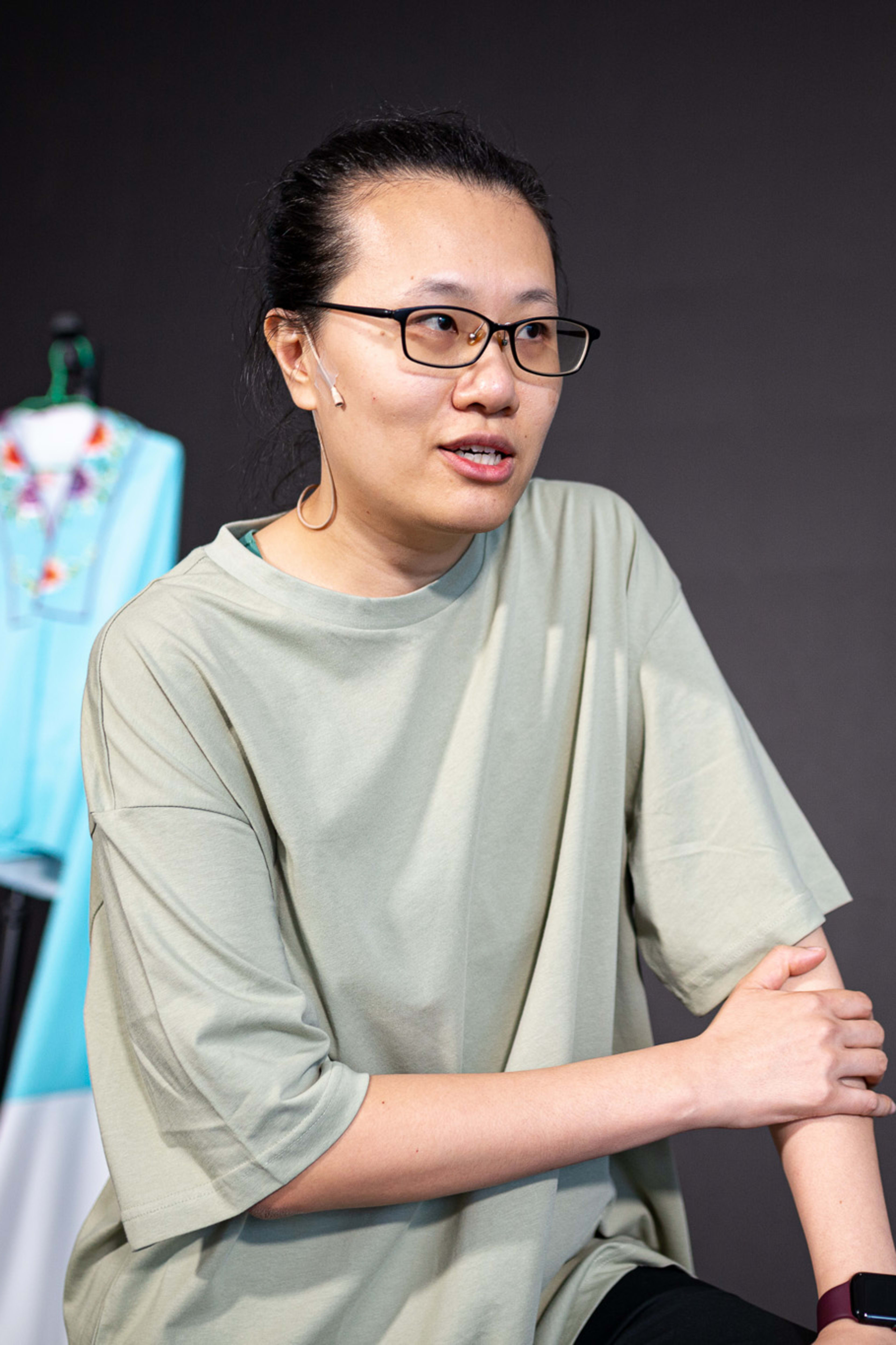 Michelle Li in a candid rehearsal shot for Rooftop Productions' contemporary theatre piece, 'Karaoke' (《卡啦OK》). She wears a casual sage green t-shirt and glasses, looking engaged and thoughtful while illuminated by soft studio lighting against a dark backdrop. A brightly colored stage costume hangs on a rack in the background. As if inviting a partner onto the stage, she says, "Jing Jing, come over here..."