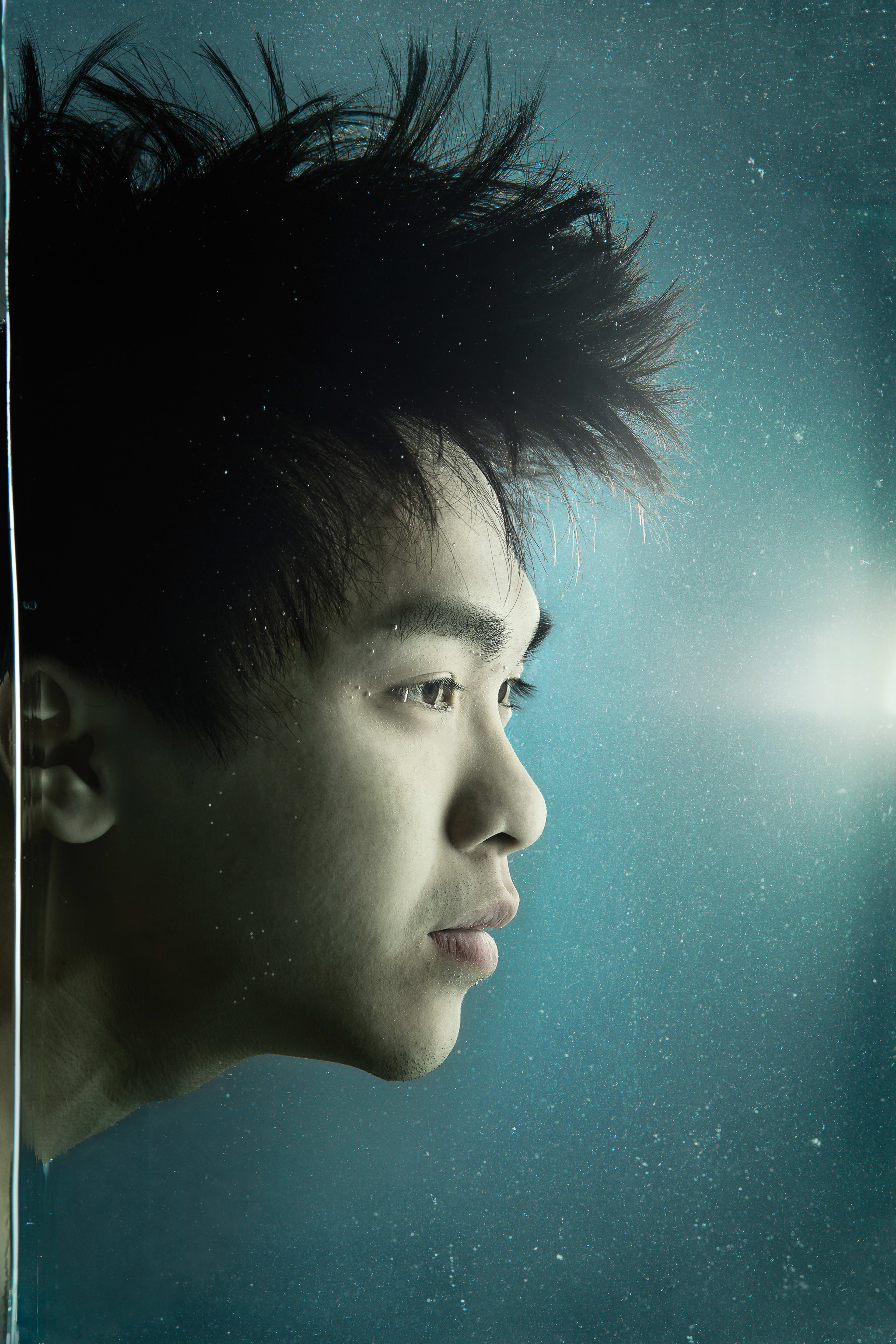 這是一張來自天台製作（Rooftop Productions）當代劇場作品《消失的海岸線》的戲劇性特寫劇照，演員吳景隆以側面示人。他置身於深青色的背景中，周圍漂浮著細小的顆粒，營造出宛如水底般的沉浸感，帶出環境消逝與記憶的主題。他神情凝重且若有所思，深刻捕捉了這部香港當代劇場表演的藝術意境。「《消失的海岸線》」