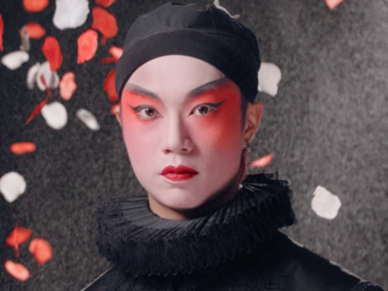 天台製作當代劇場作品《曲解莎劇》（History Play）主視覺海報。演員梁俊仁飾演 Rizzly（Southampton伯爵），他面容沉靜，化著大膽且具前衛感的舞台妝容，深紅色眼影與紅唇對比強烈。他身穿精緻的黑色伊莉莎白風格服裝，頸部佩戴黑色拉夫領（ruff collar）。背景為充滿質感的深色調，紅、白玫瑰花瓣在四周飄落，營造出強烈的戲劇氛圍與香港當代劇場的獨特美學。《曲解莎劇》讀劇演出。