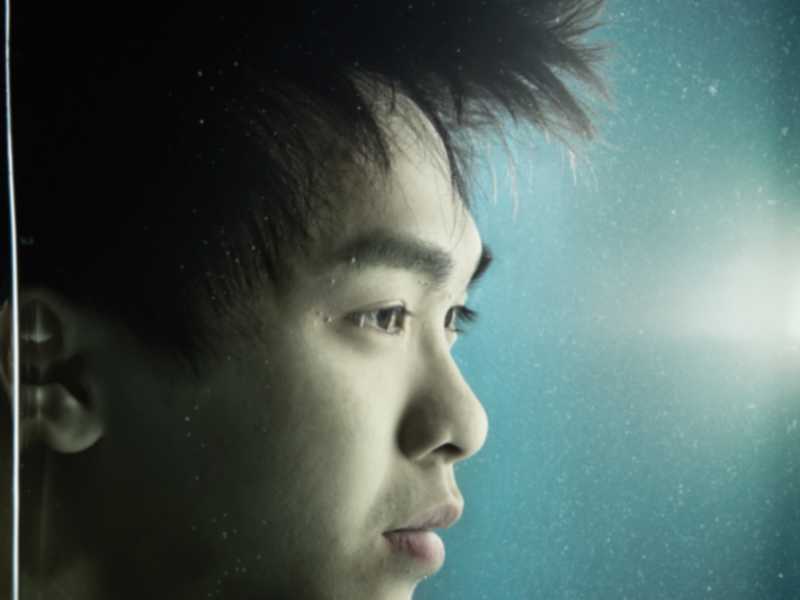 這是一張來自天台製作（Rooftop Productions）當代劇場作品《消失的海岸線》的戲劇性特寫劇照，演員吳景隆以側面示人。他置身於深青色的背景中，周圍漂浮著細小的顆粒，營造出宛如水底般的沉浸感，帶出環境消逝與記憶的主題。他神情凝重且若有所思，深刻捕捉了這部香港當代劇場表演的藝術意境。「《消失的海岸線》」