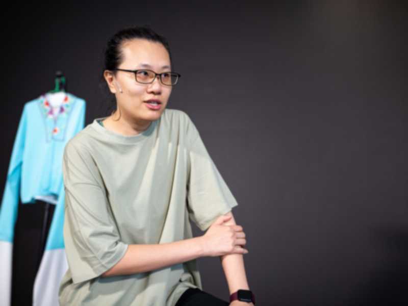 Michelle Li in a candid rehearsal shot for Rooftop Productions' contemporary theatre piece, 'Karaoke' (《卡啦OK》). She wears a casual sage green t-shirt and glasses, looking engaged and thoughtful while illuminated by soft studio lighting against a dark backdrop. A brightly colored stage costume hangs on a rack in the background. As if inviting a partner onto the stage, she says, "Jing Jing, come over here..."