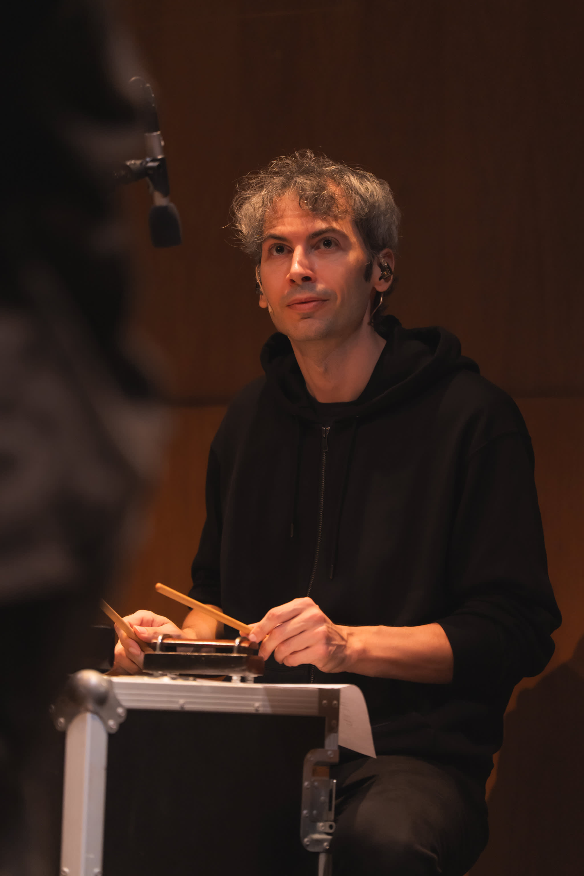 音樂家艾浩家（Ivor Houlker）身穿黑色連帽衫，佩戴入耳式耳機，在天台製作（Rooftop Productions）的現代劇場作品《曲解莎劇》中進行現場演奏。他正專注地手持木槌，在溫暖昏黃的舞台燈光與木質背景前表演。Ivor。