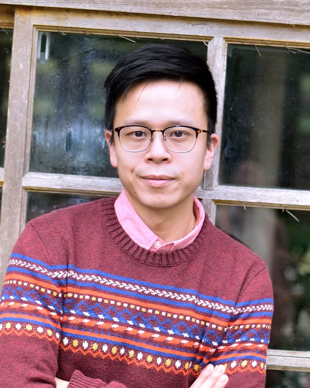 A professional headshot of Hong Kong theatre actor Hollis Ngai, captured in a soft, natural light against a grid-like wooden window frame background. He wears a maroon patterned sweater over a light pink collared shirt, projecting a thoughtful and approachable demeanor characteristic of the contemporary performers at Rooftop Productions (天台製作). A headshot of Hollis Ngai.