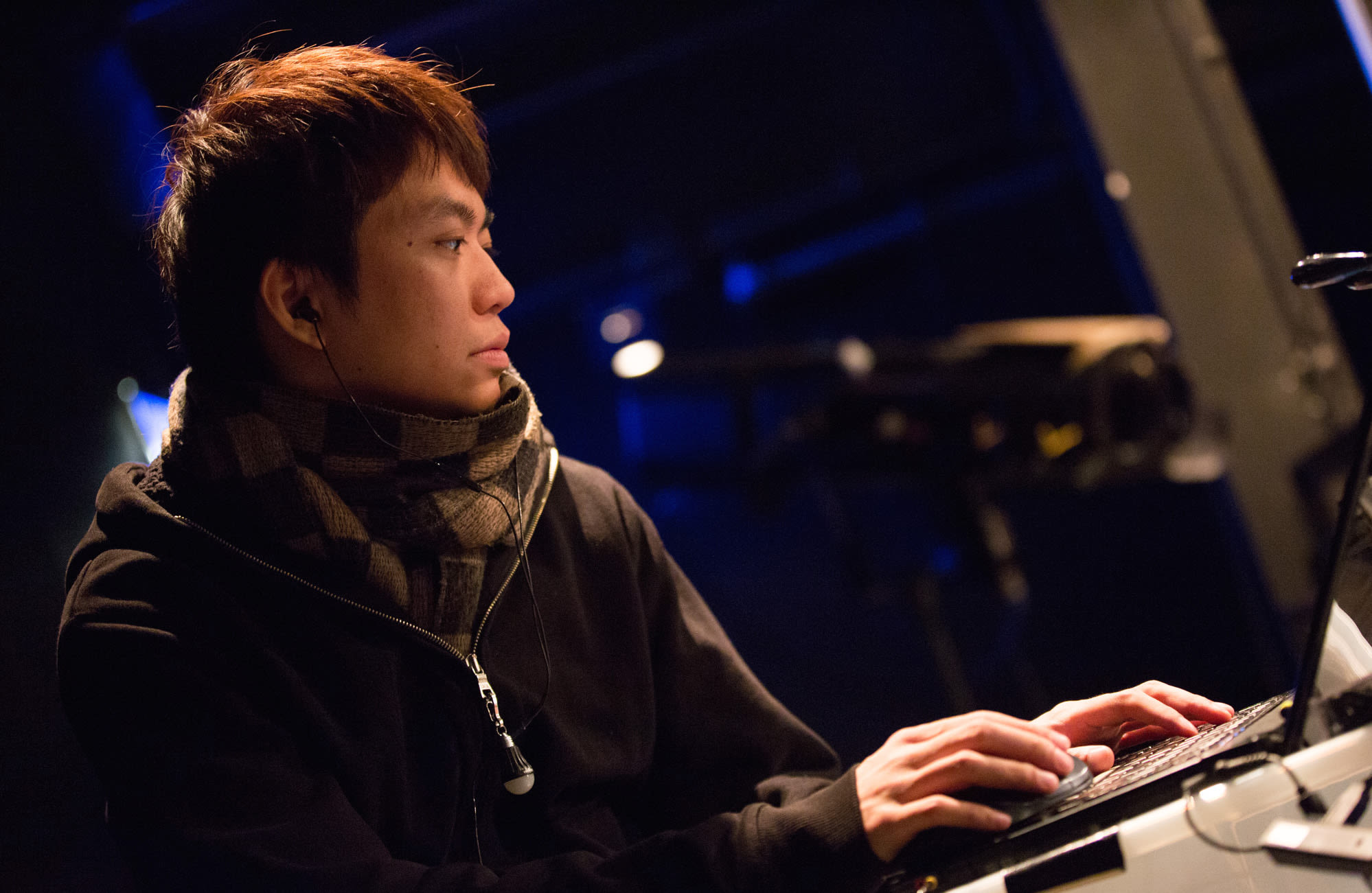 歐陽翰奇（Au Yeung Hon Ki）於天台製作（Rooftop Productions）當代香港劇場排練期間的側拍特寫。照片中他身穿深色連帽衫與格紋圍巾，正專注地注視著舞台控制台上的筆記型電腦螢幕，周圍環繞著劇場特有的冷調藍光。這張照片捕捉了演出前的技術排練時刻。個人特寫照。