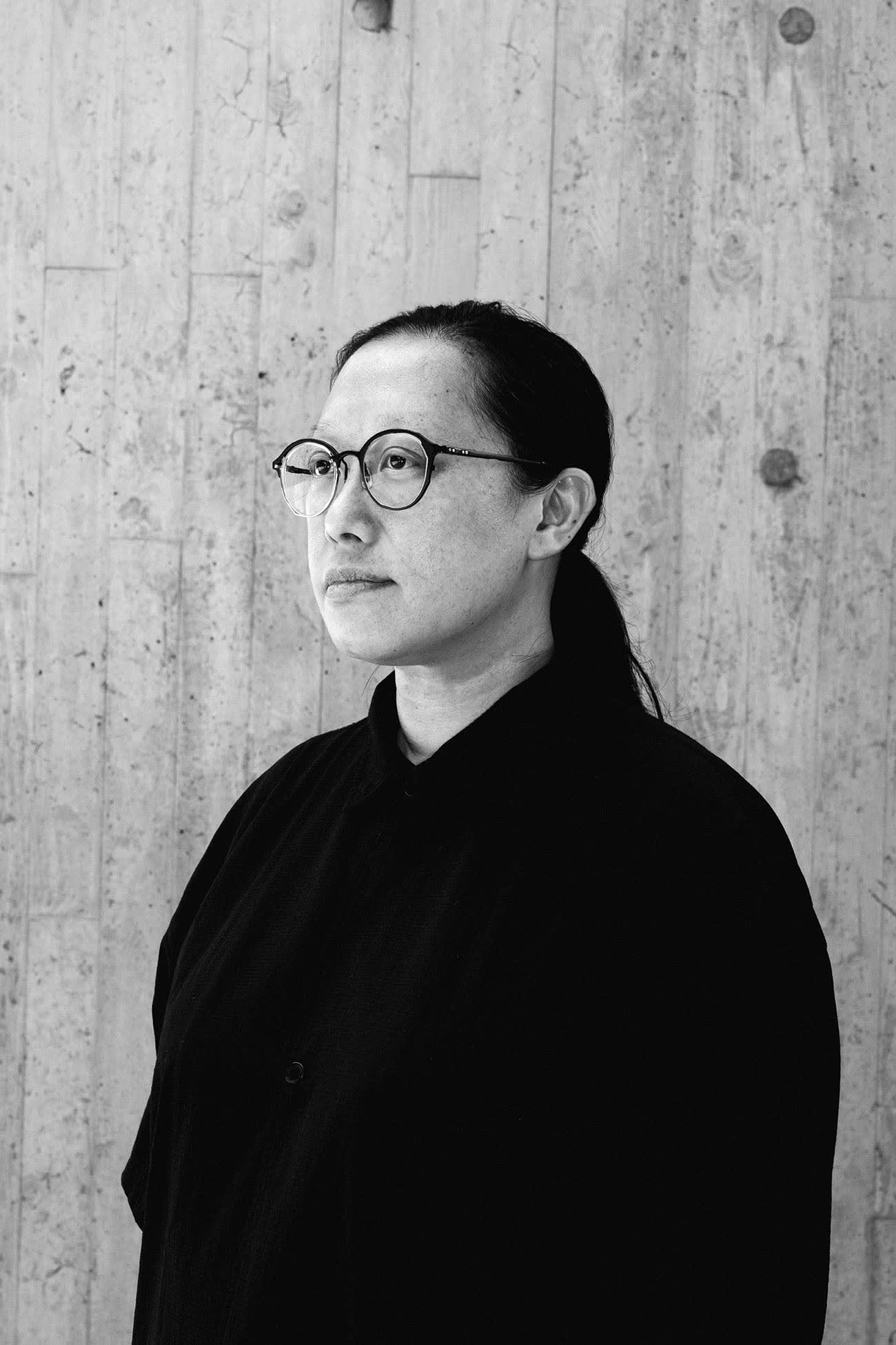 這是一張演員李筱怡（Bobo Lee）的黑白定裝照，背景為充滿香港當代劇場質感的水泥牆面。李筱怡配戴深色膠框眼鏡，身穿黑色襯衫，側身望向遠方，展現出天台製作（Rooftop Productions）極簡且具實驗性的舞台美學風格。定裝照。