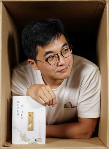 天台製作（Rooftop Productions）演員陳冠宏的劇照。照片中他身處一個紙箱內，雙手撐在箱緣，手持書籍《就有多自由，你就有多自律》。溫暖的攝影棚燈光映照著他若有所思的神情，呈現出香港當代劇場獨特的實驗感與親密氛圍。陳冠宏。