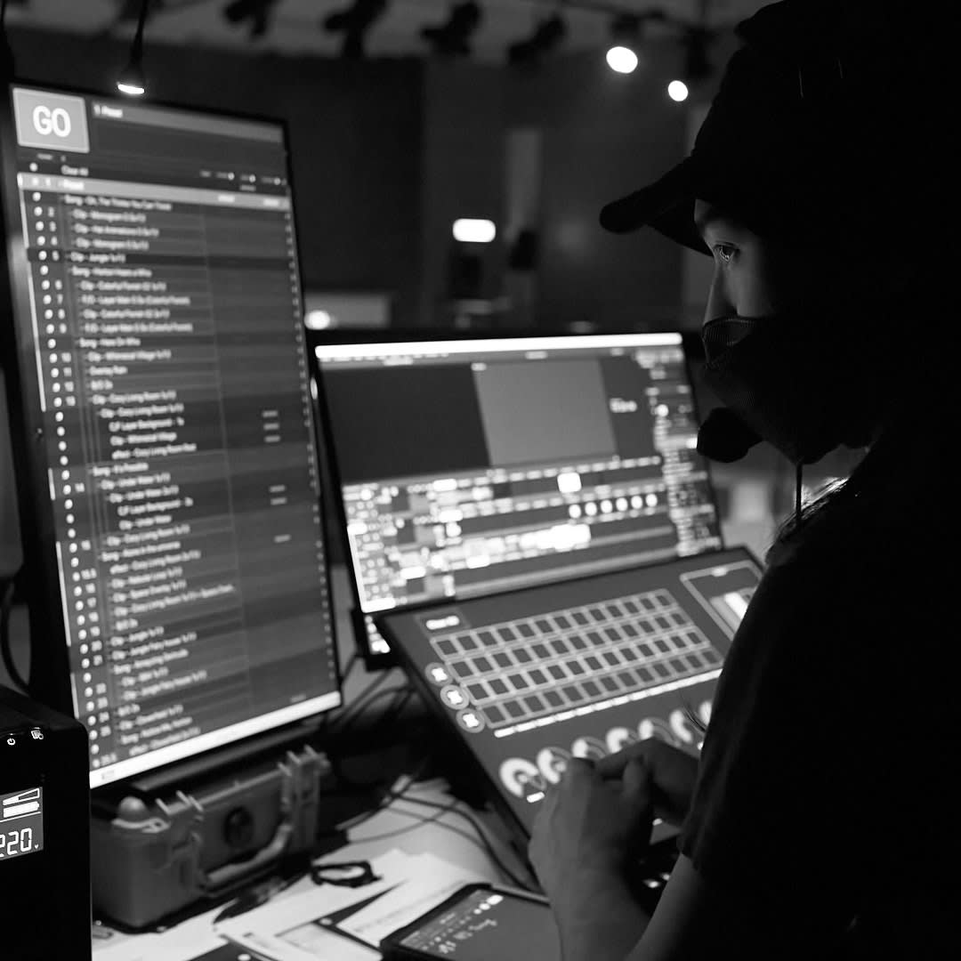 A black-and-white, atmospheric shot of performer Chan Tsz Ho Calvin focused at a workstation, reflecting the technical precision of Rooftop Productions' (天台製作) contemporary theatre practice. He is silhouetted against the bright, detailed displays of media server software and a lighting console, capturing a quiet, professional moment of behind-the-scenes production work.