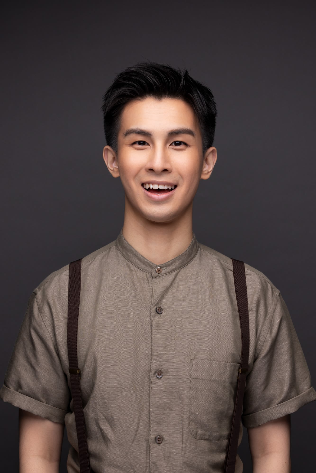 Professional headshot of actor Cheung Cheuk-hang, captured against a clean, neutral grey studio backdrop. He is wearing a minimalist, taupe-colored short-sleeved button-down shirt paired with brown suspenders, reflecting the contemporary aesthetic often seen in Rooftop Productions (天台製作) theatre performances. He looks directly at the camera with a warm, friendly smile, conveying a sense of artistic readiness and poise typical of a Hong Kong stage performer.