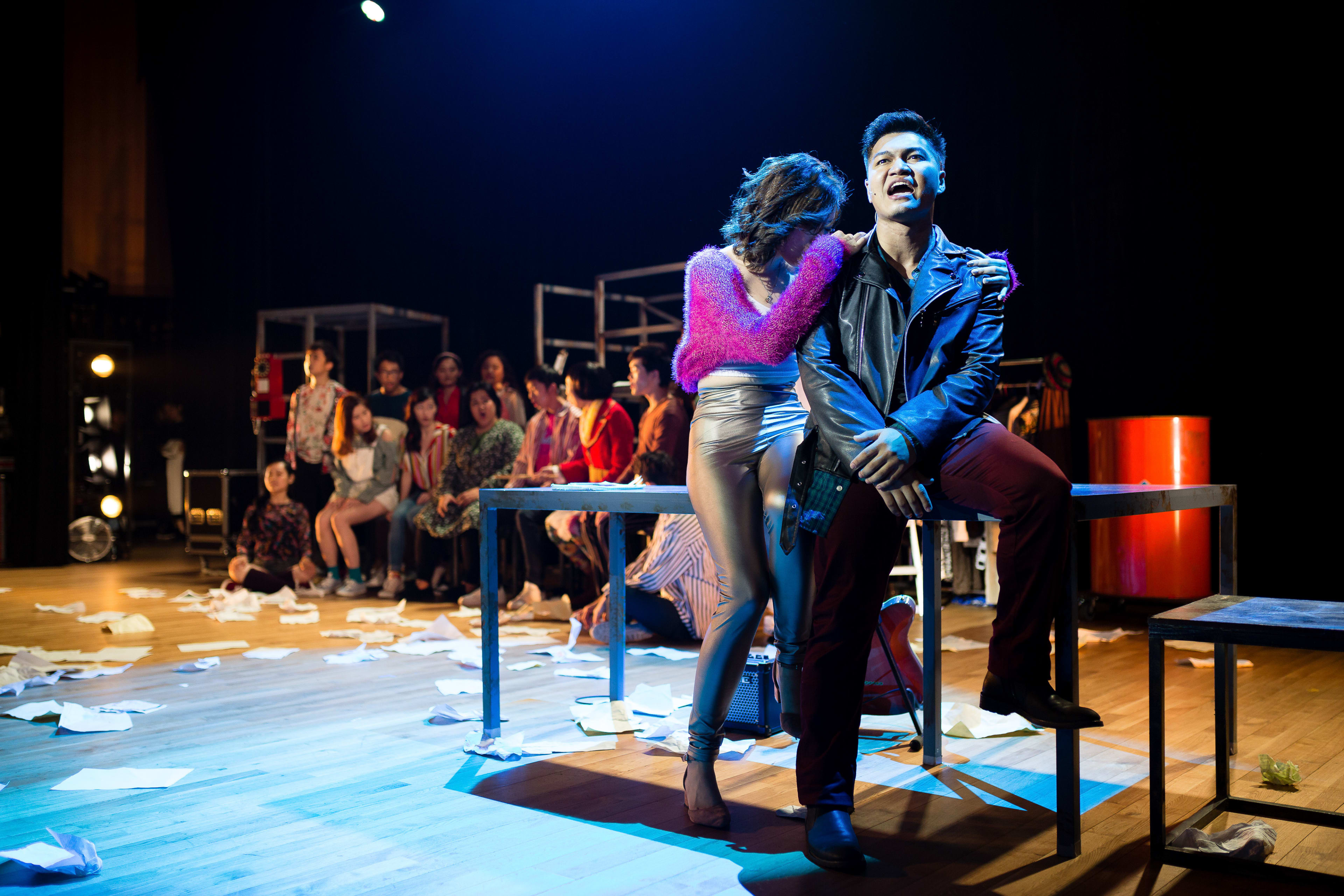 A dramatic stage scene from the Hong Kong production of the rock musical 'RENT' by Rooftop Productions (天台製作). In a dimly lit, atmospheric theatre setting strewn with scattered papers, an actor portraying a character sits on a table while an actress in a fuzzy pink sweater leans against him, expressing intense emotion. The staging reflects the raw, gritty energy of contemporary HK theatre, with a live ensemble visible in the blurred background. As the characters share a poignant moment, the male lead sings, 'RENT'.