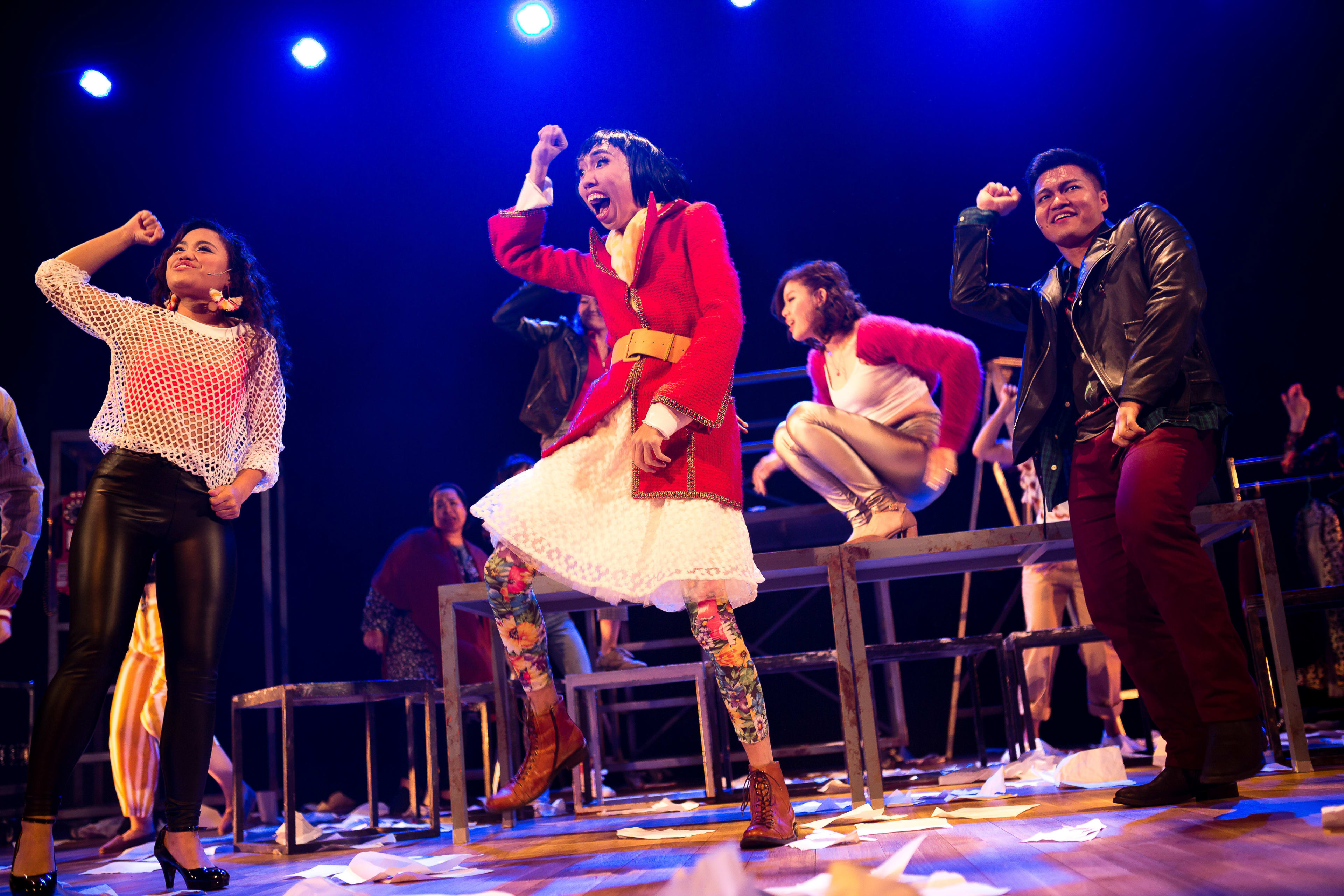 In this dynamic scene from Rooftop Productions' staging of RENT, five performers express high-energy emotion on a stage strewn with scattered paper. Center stage, an actor stands tall in a vibrant red coat and patterned leggings, cheering with arms raised. To the left, a performer in a white mesh top and black pants poses with confidence, while another performer leaps on a raised platform behind them. To the right, an actor in a leather jacket smiles broadly, capturing the spirit of contemporary Hong Kong theatre. As the cast brings the iconic musical to life, they sing, "RENT."