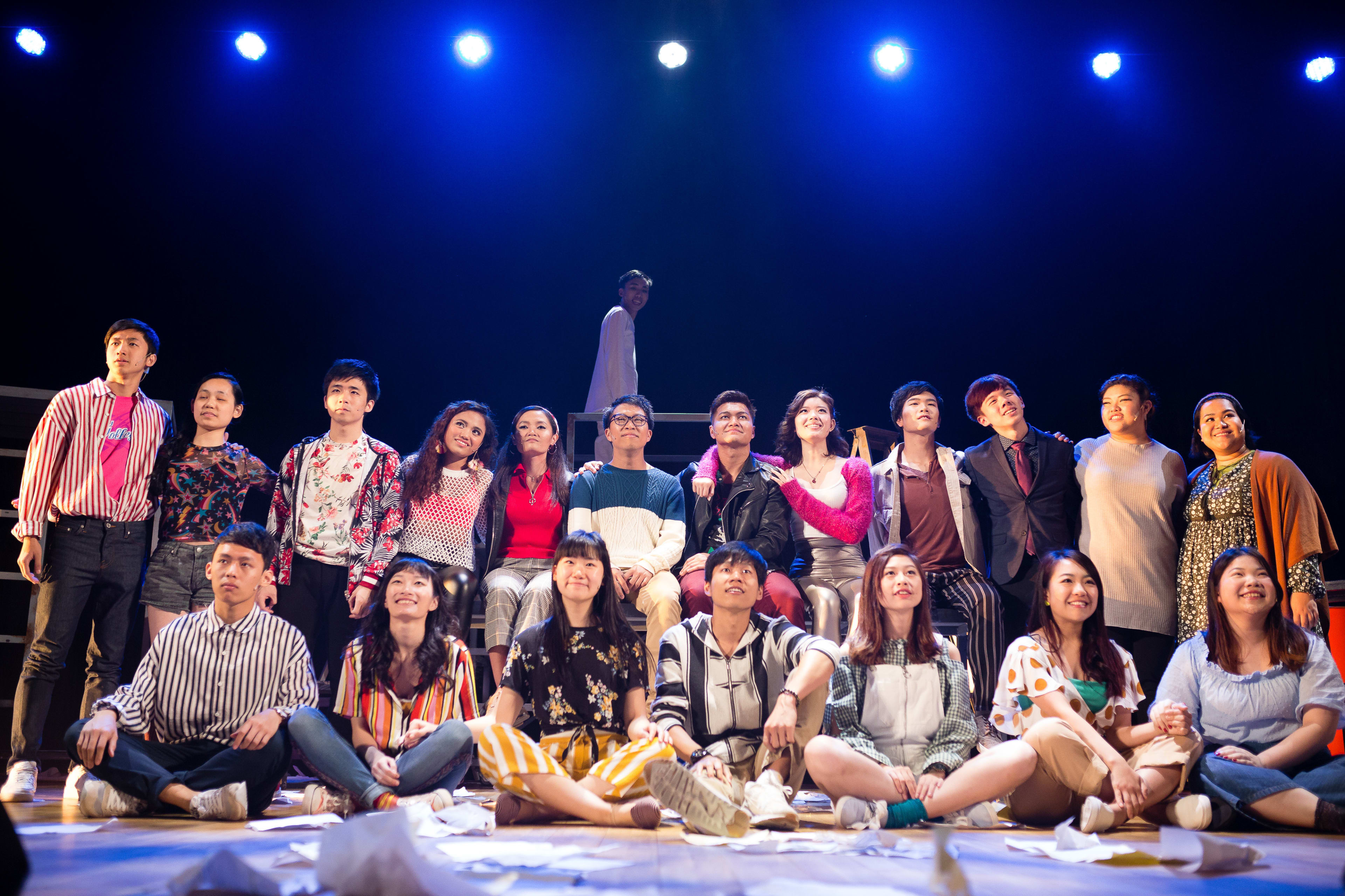 A wide-angle shot of a diverse ensemble cast performing in Rooftop Productions' contemporary staging of the rock musical 'RENT'. The actors are arranged in a dynamic composition, with some sitting on the stage floor and others standing behind them, all looking upward with expressions of hope and determination under bright, theatrical spotlights. The setting evokes a raw, industrial urban atmosphere common in Hong Kong's independent theatre scene. 'RENT'