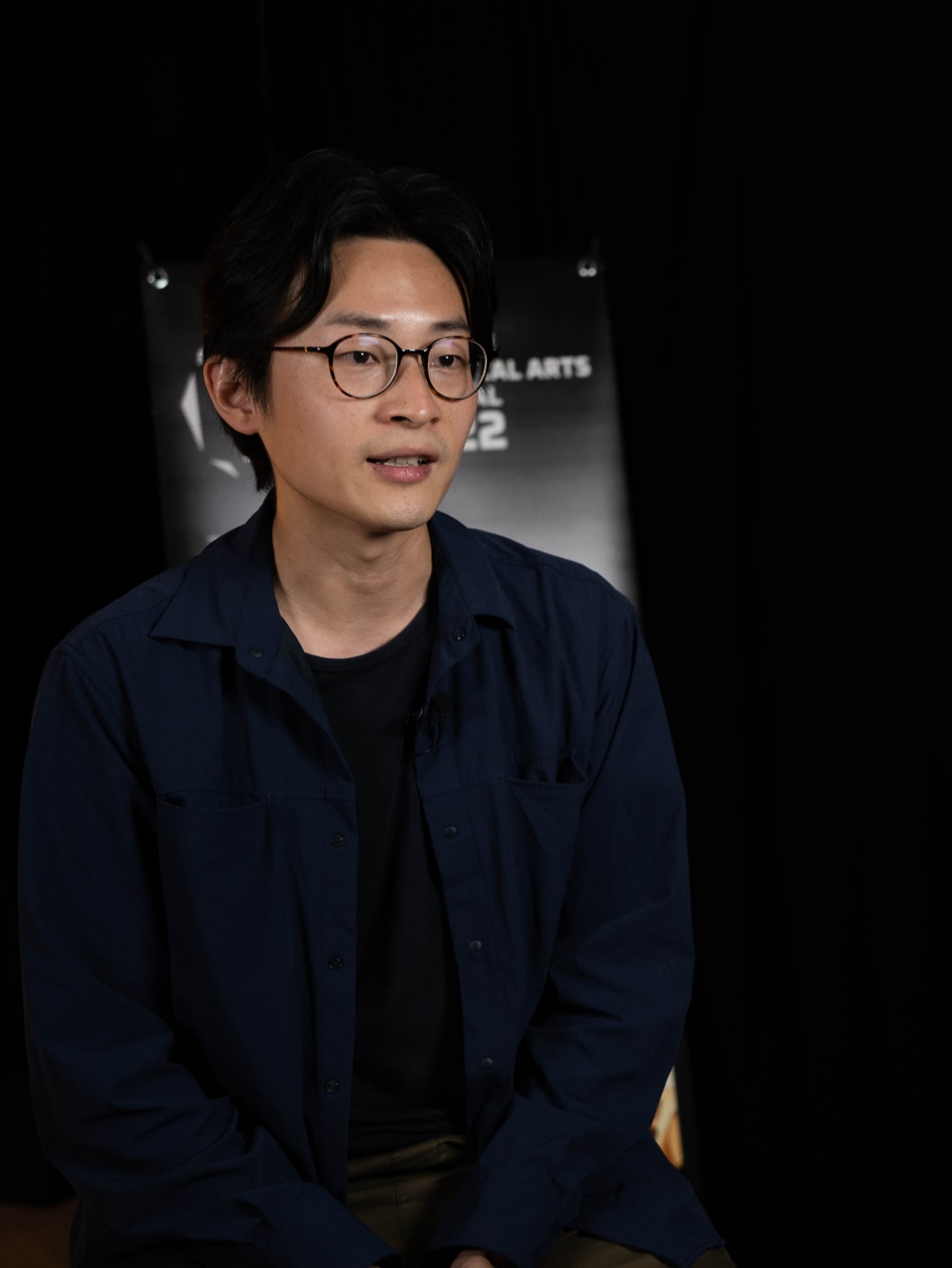 A studio headshot of Hong Kong theatre actor Fong Hiu-tan, who is wearing round-rimmed glasses and a dark blue button-down shirt. He is captured in a soft, focused light against a dark background, reflecting the professional and artistic atmosphere typical of Rooftop Productions (天台製作) theatre work. A headshot.