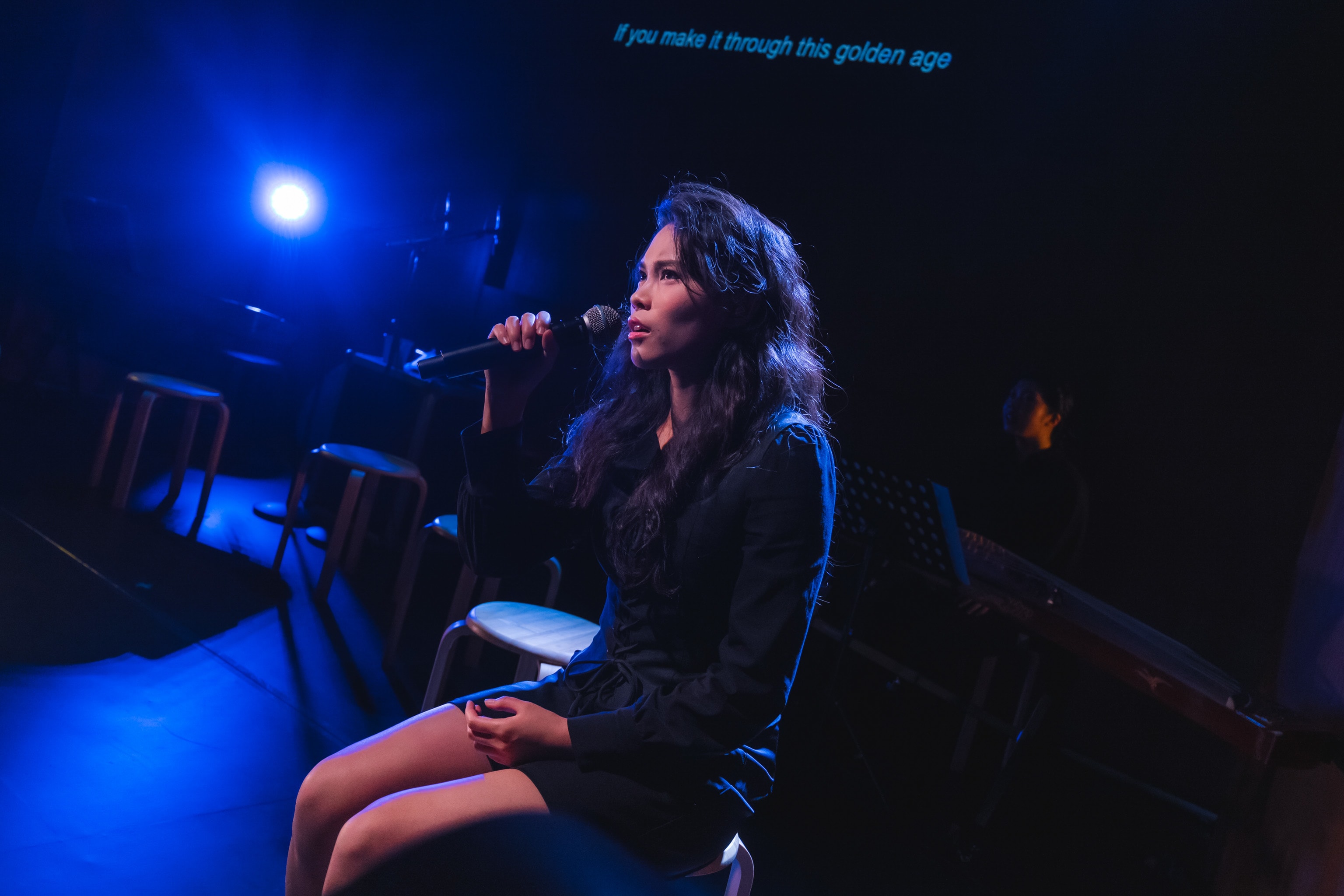 錢穎嘉在天台製作的當代香港舞台劇《曲解莎劇》中飾演 Aemilia Lanier。舞台燈光呈現戲劇性的藍色，錢穎嘉坐在高腳凳上，手持麥克風深情演唱，情感投入。舞台環境簡約且黑暗，陰影處隱約可見樂器。她唱道：「日日盡力活在大時代。」