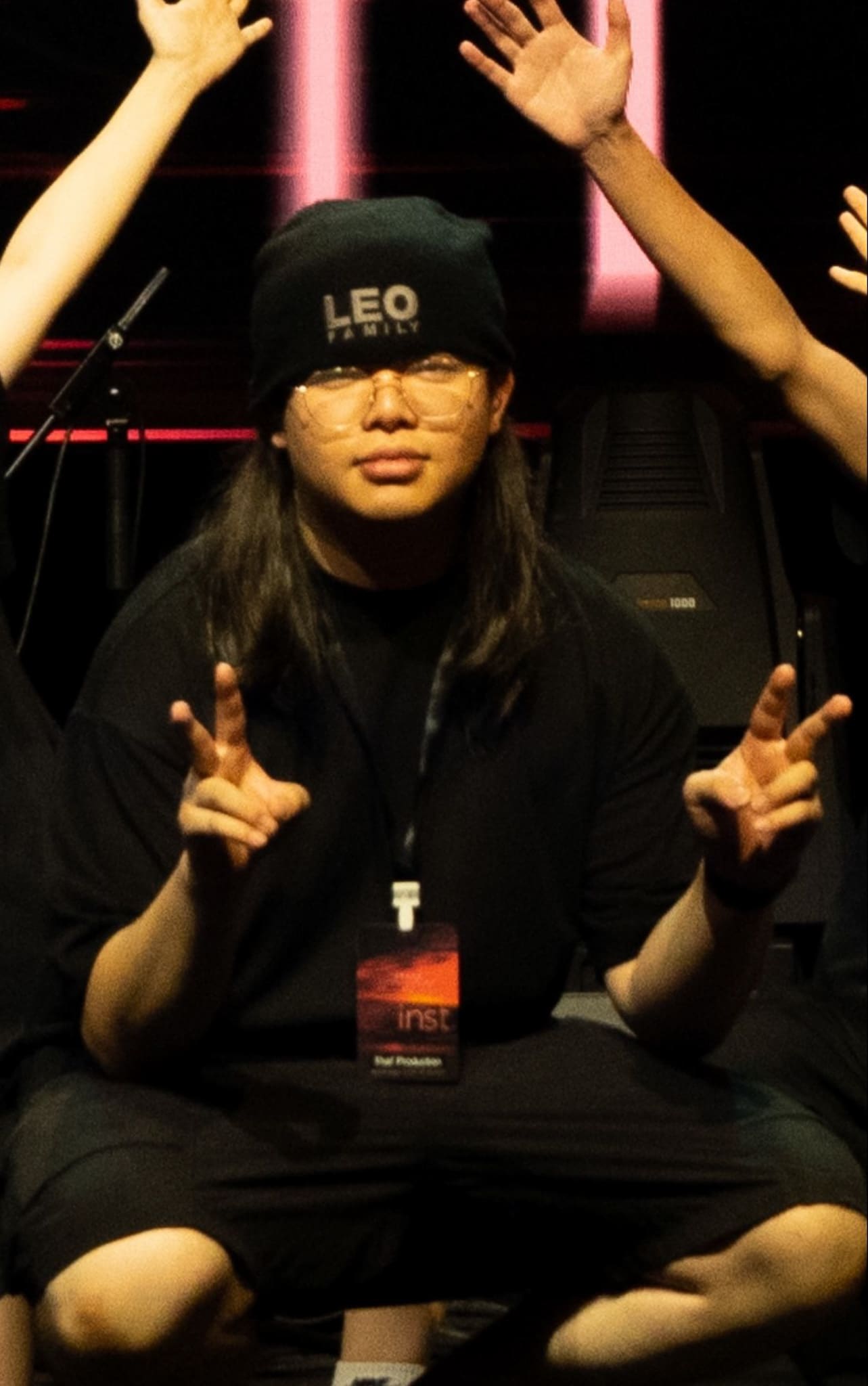 天台製作 (Rooftop Productions) 成員蘇浩文 (Homan So) 在舞台上蹲坐，戴著印有「LEO FAMILY」字樣的黑色冷帽與眼鏡，雙手比出 V 字手勢。現場燈光以充滿現代感的紅色作為背景，展現出香港當代劇場排練或演出現場的真實氛圍。劇照。