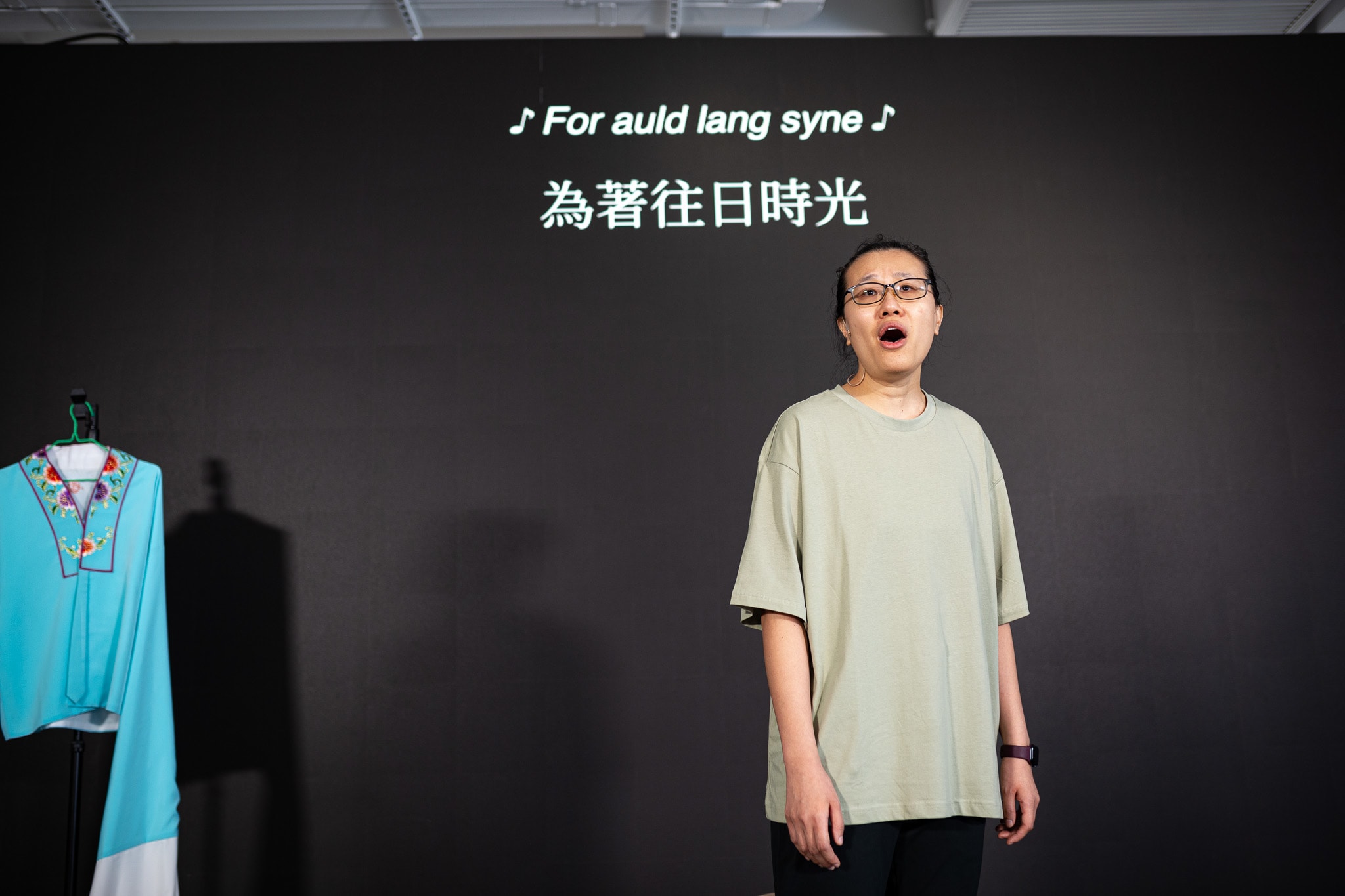 在天台製作 (Rooftop Productions) 當代香港劇場作品《卡啦OK》的排練中，演員李婉晶站在極簡主義的黑色背景舞台前。她身穿淺色T恤，戴著眼鏡，神情投入地歌唱，舞台左側懸掛著一件亮藍色的演出服。背景上方投影著歌詞「為著往日時光」。李婉晶透過歌聲表達了劇中的情感：「為著往日時光」。