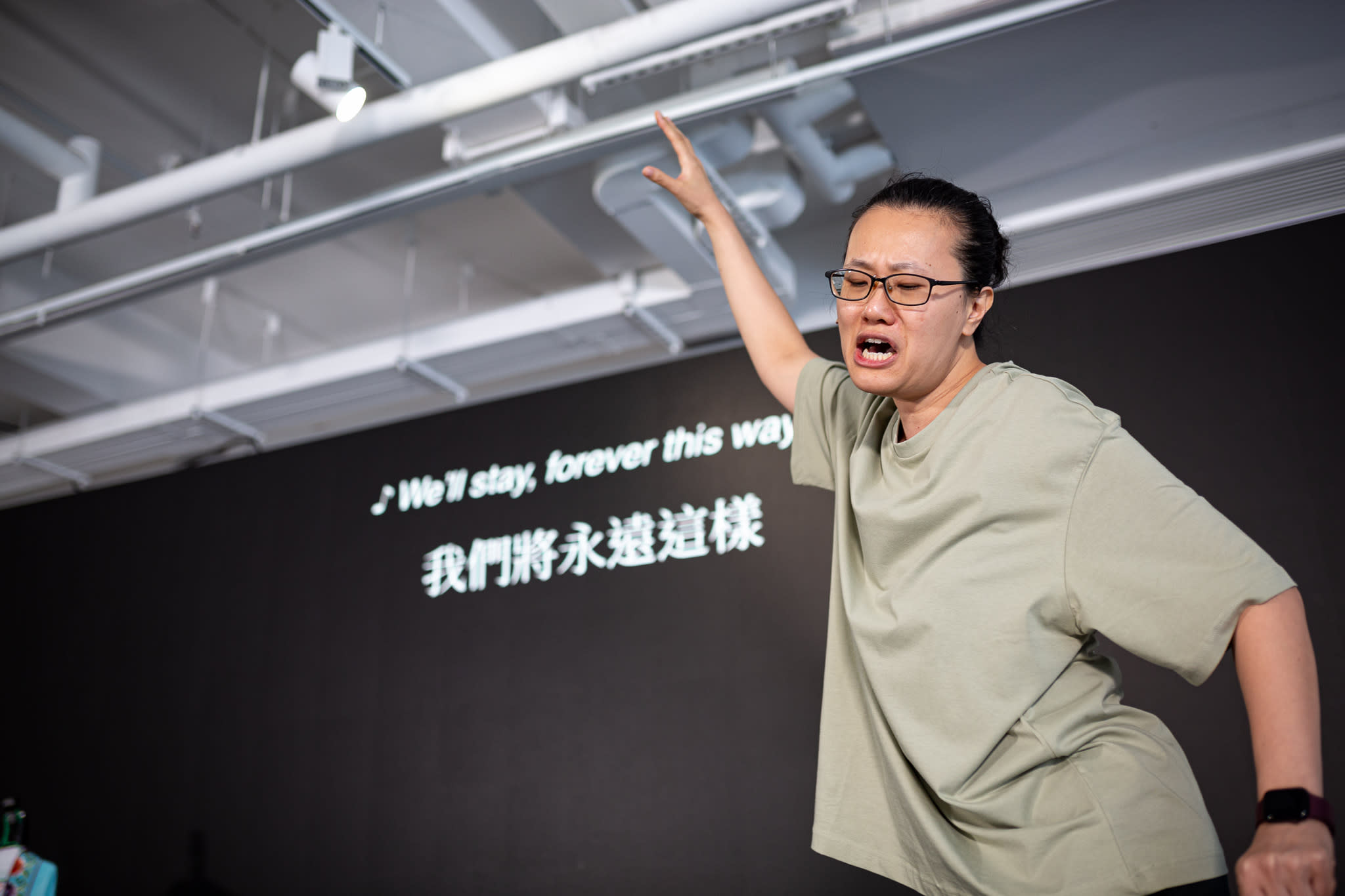 李婉晶在天台製作的當代劇場作品《卡啦OK》排練中演出。在攝影棚燈光的映照下，她神情激昂，手臂向外伸展，展現出強烈的情感張力，身後的黑色牆面上投射著演出歌詞。她正深情唱出：「我們將永遠這樣。」
