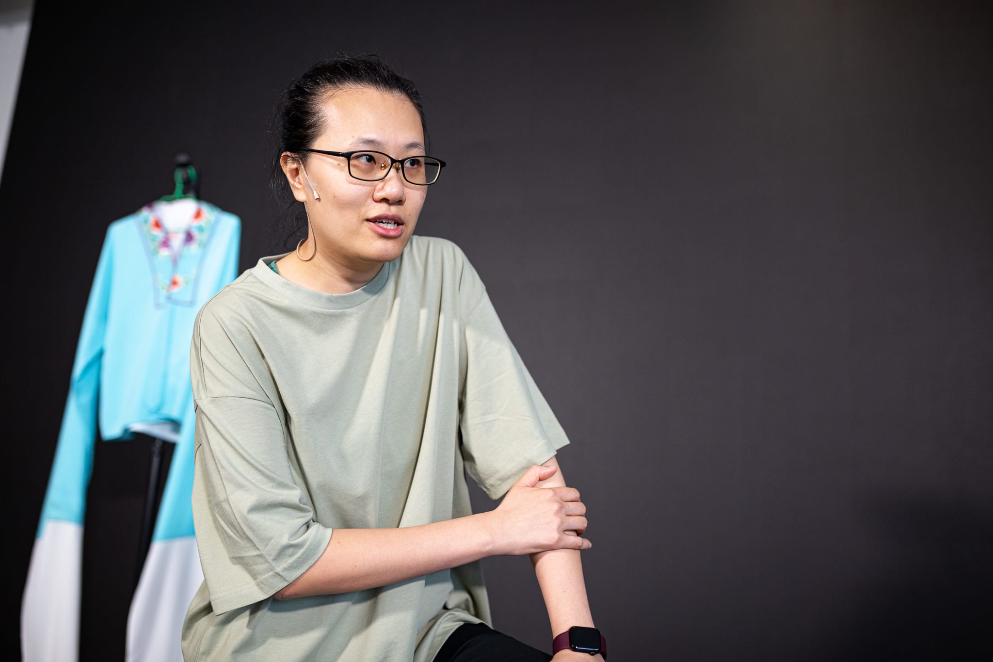 李婉晶在天台製作當代劇場作品《卡啦OK》的排練場照。她身穿休閒鼠尾草綠色T恤及佩戴眼鏡，在暗色背景的柔和燈光下顯得專注而深思。背景衣架上掛著色彩鮮豔的戲服。她彷彿正在邀請夥伴走上舞台，說道：「晶晶，過來啊。」