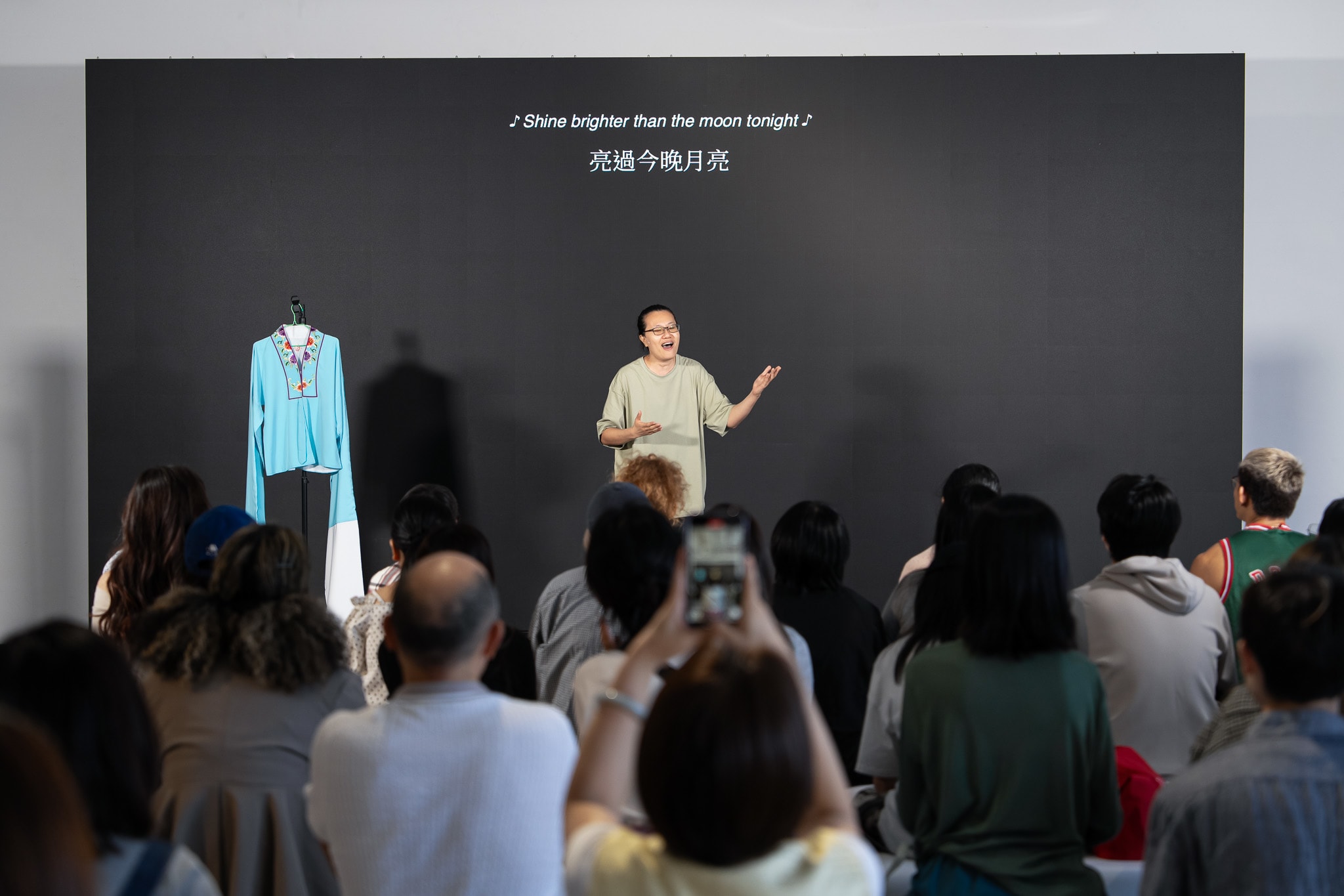 在天台製作的當代香港劇場作品《卡啦OK》中，演員李婉晶站在極簡的舞台上，背靠黑色的背景牆，投入地演唱並配合身體語言。她的左側懸掛著一件衣物，在燈光下顯得格外突出。觀眾席視角捕捉了演出現場，前排觀眾正舉起手機記錄這一瞬間。背景牆上投射著劇中台詞：「亮過今晚月亮」