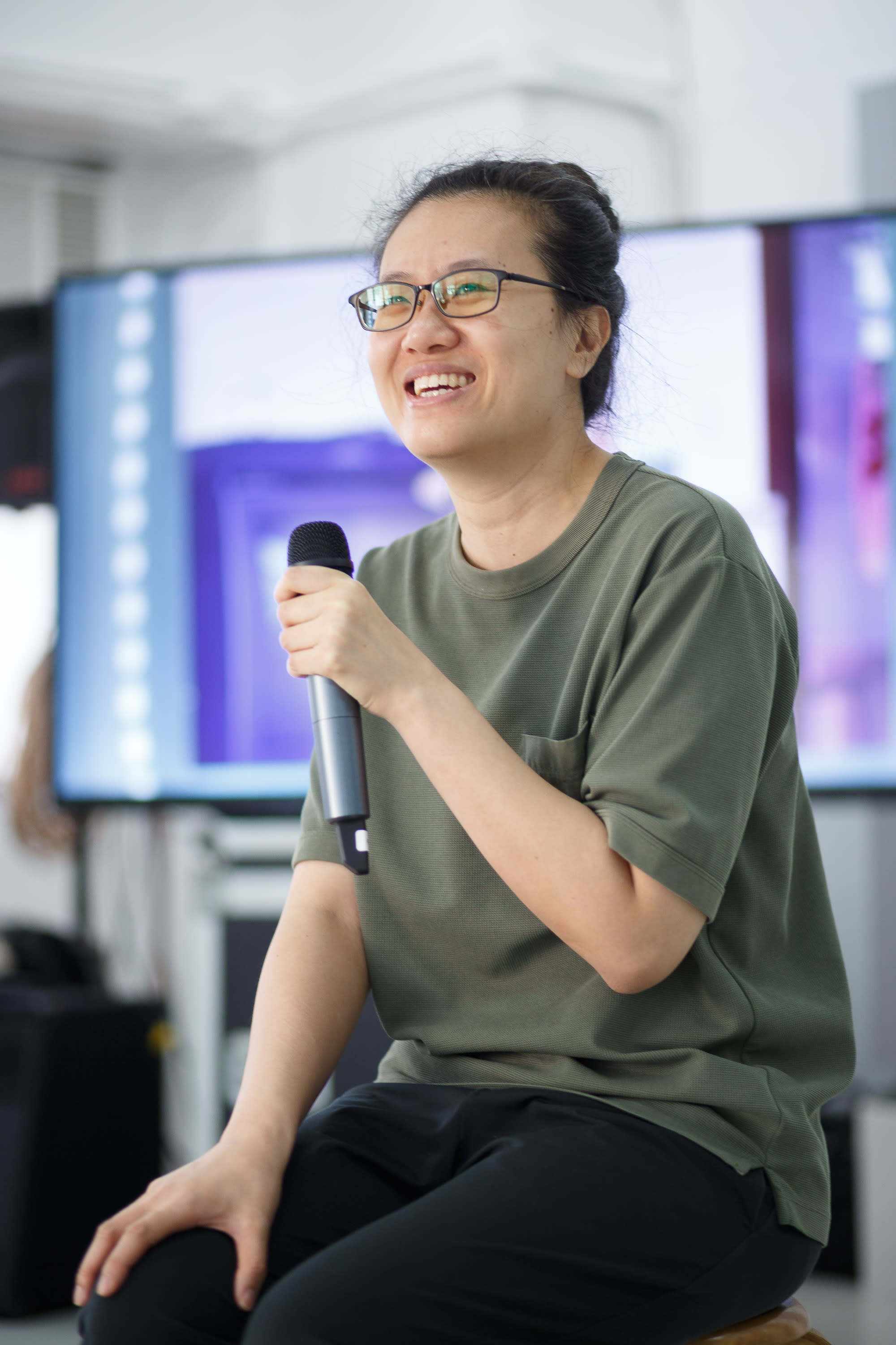 演員李婉晶在天台製作的當代劇場作品《卡啦OK》排練過程中，手持麥克風微笑。她身穿休閒橄欖綠色上衣與眼鏡，畫面記錄了排練室中真摯且充滿張力的創作瞬間。《卡啦OK》排練