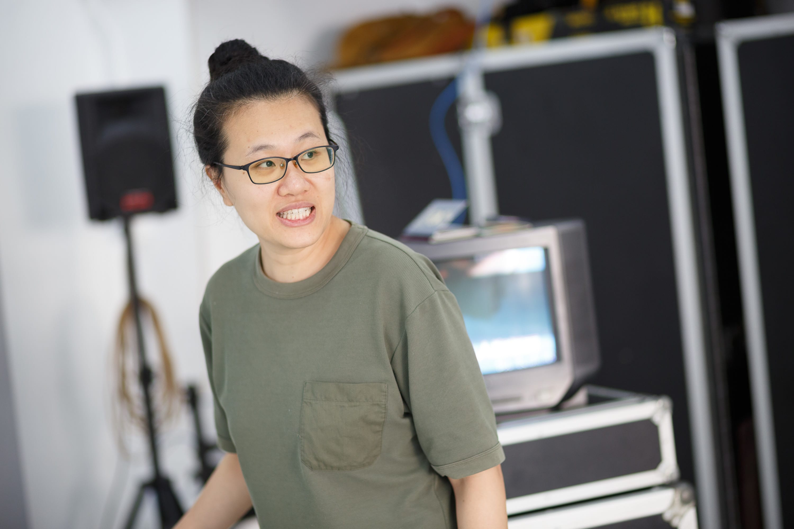 Performer Michelle Li (李婉晶) captured in a candid moment during the production rehearsals for the contemporary Hong Kong theatre play 'Karaoke' (《卡啦OK》). Li wears a casual olive-green t-shirt with her hair tied up, her expression focused and animated as she engages in a scene. The staging setup in the background includes technical equipment, speakers, and a vintage television monitor, reflecting the raw, behind-the-scenes atmosphere of the rehearsal space. 'Karaoke rehearsals'