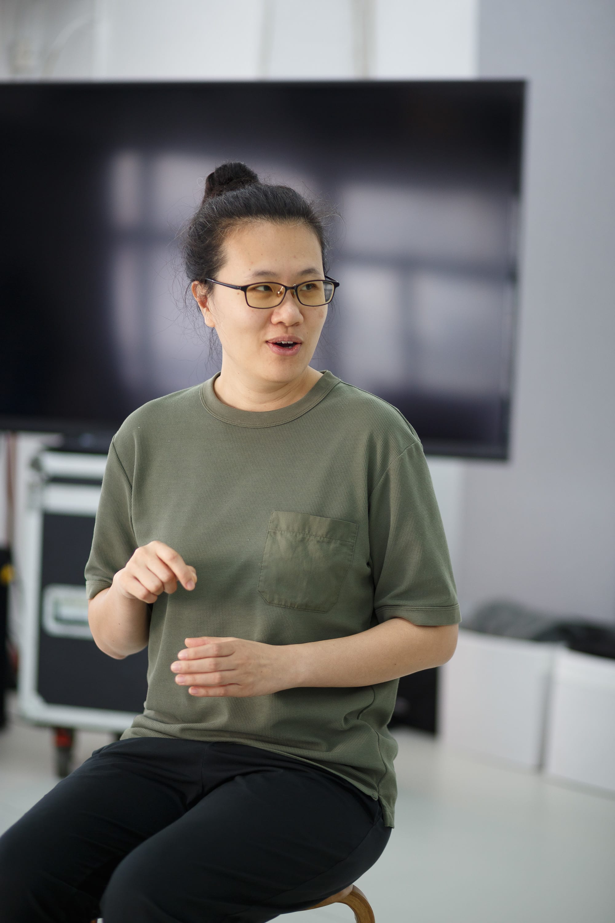 Performer Michelle Li is captured in a focused moment during a rehearsal for the contemporary Hong Kong stage production 'Karaoke' (《卡啦OK》). She wears a casual olive green t-shirt and glasses, gesturing with her hands as if in conversation or mid-performance, with the blurred background of a rehearsal studio and a large screen behind her. 'Karaoke rehearsals'