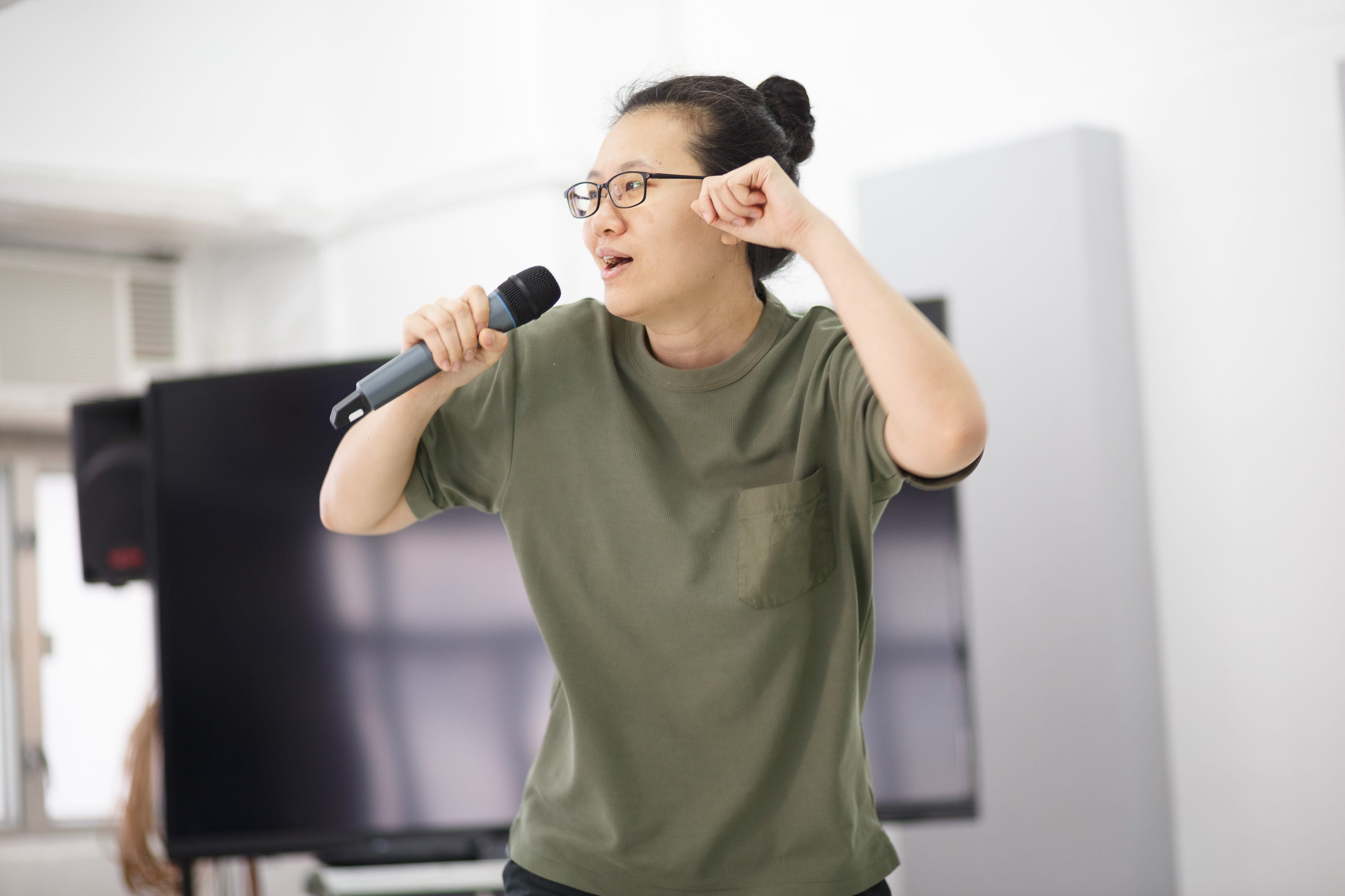 Performer Michelle Li holds a microphone and gestures with her other hand, captured during an intense rehearsal for Rooftop Productions' contemporary Hong Kong theatre piece, 'Karaoke'. Her focused expression highlights the raw energy of the performance. 'Karaoke rehearsals'