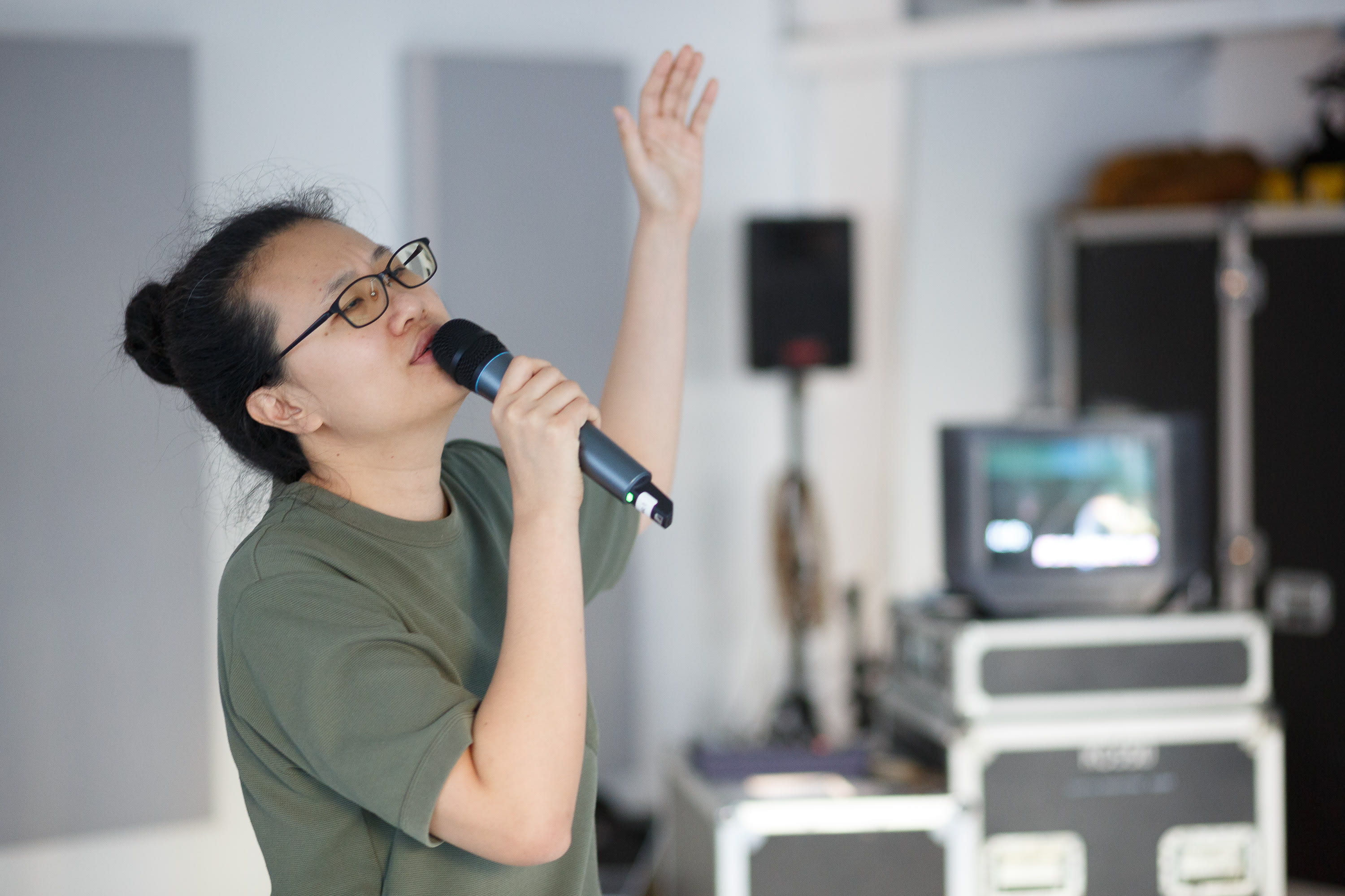李婉晶正在為天台製作的當代劇場作品《卡啦OK》進行排練。相片以中景拍攝，戴著眼鏡的李婉晶身穿簡約T恤，情感飽滿地對著麥克風演唱，左手高舉向天。背景呈現了排練室常見的設備，包括顯示器、揚聲器腳架和器材箱，展現了這部戲劇創作背後的真實排練氛圍。《卡啦OK》排練