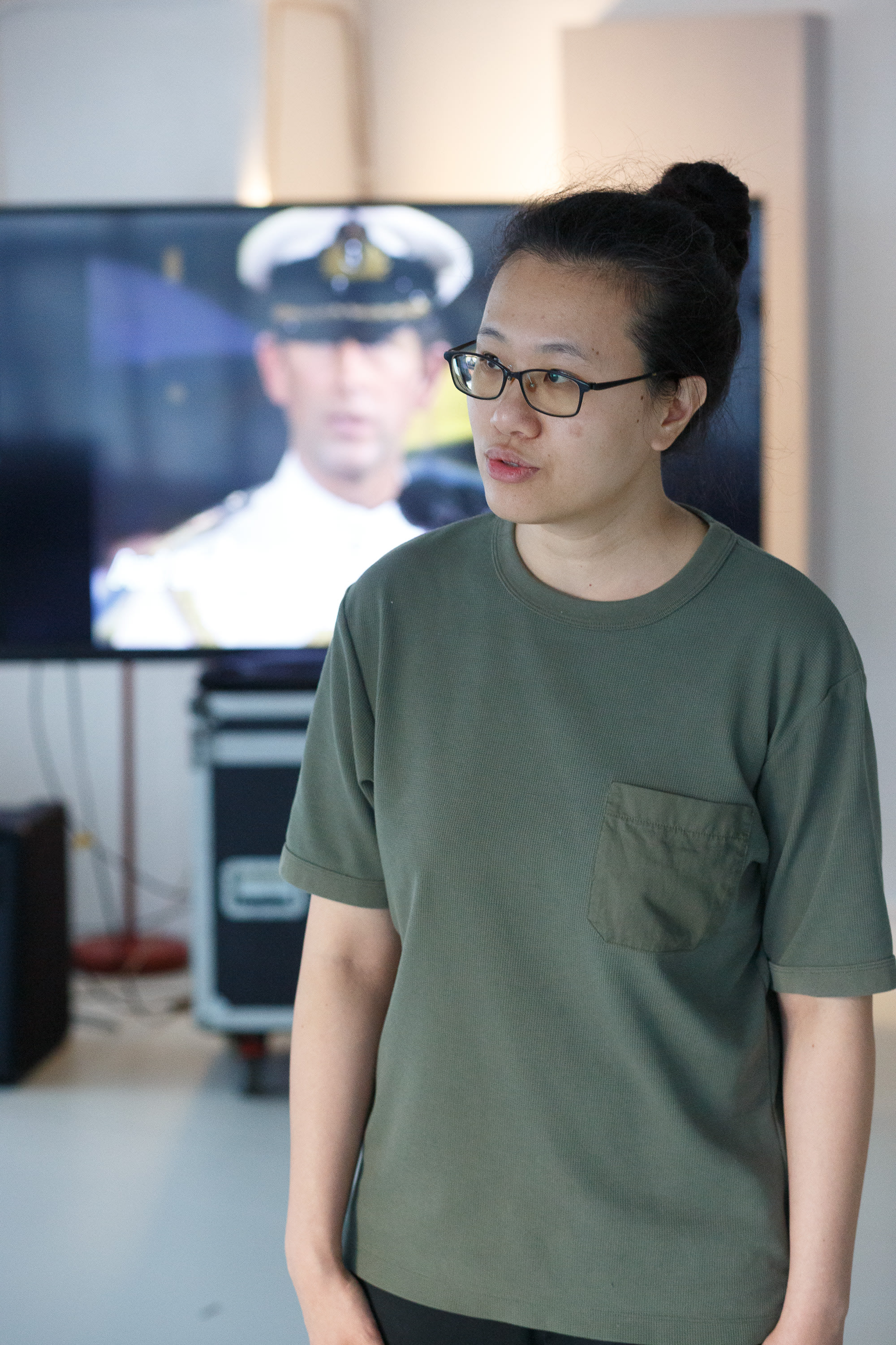 在天台製作 (Rooftop Productions) 當代劇場作品《卡啦OK》的排練現場，演員李婉晶（Michelle Li）佩戴眼鏡，身穿橄欖綠色口袋T恤，將頭髮盤起，神情專注地進行表演排練。背景中的大型螢幕投影著一位身穿海軍軍官制服的人物影像，為這場香港舞台演出增添了一層關於記憶或身分認同的敘事意涵。《卡啦OK》排練