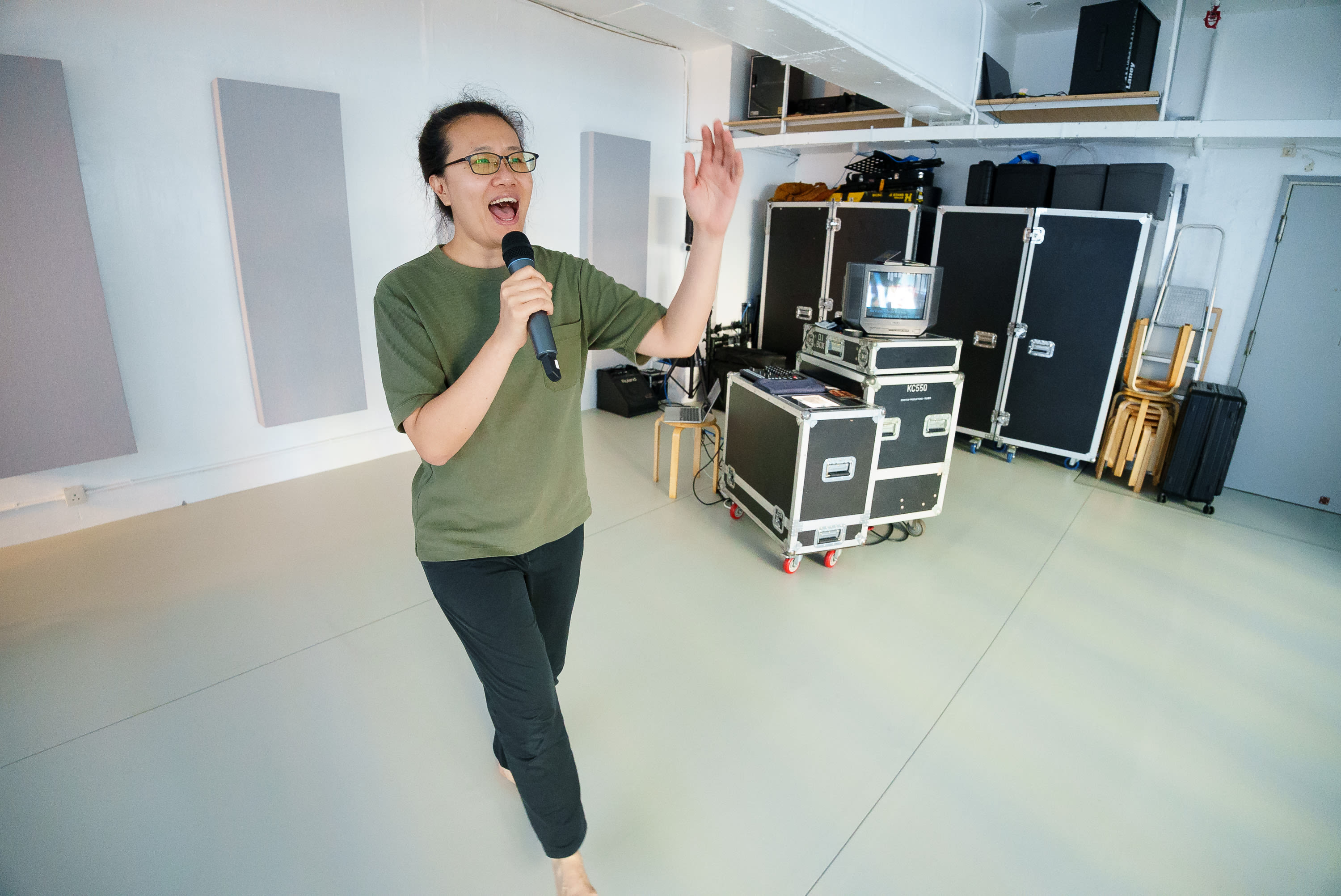 Michelle Li performing during a rehearsal for the Rooftop Productions contemporary theatre show 'Karaoke'. She is captured in mid-action, holding a microphone in one hand and gesturing with the other while singing with an expressive, passionate facial expression. The minimalist studio space features soundproofing panels and stage equipment, highlighting the raw energy of the performance. 'Karaoke rehearsals'