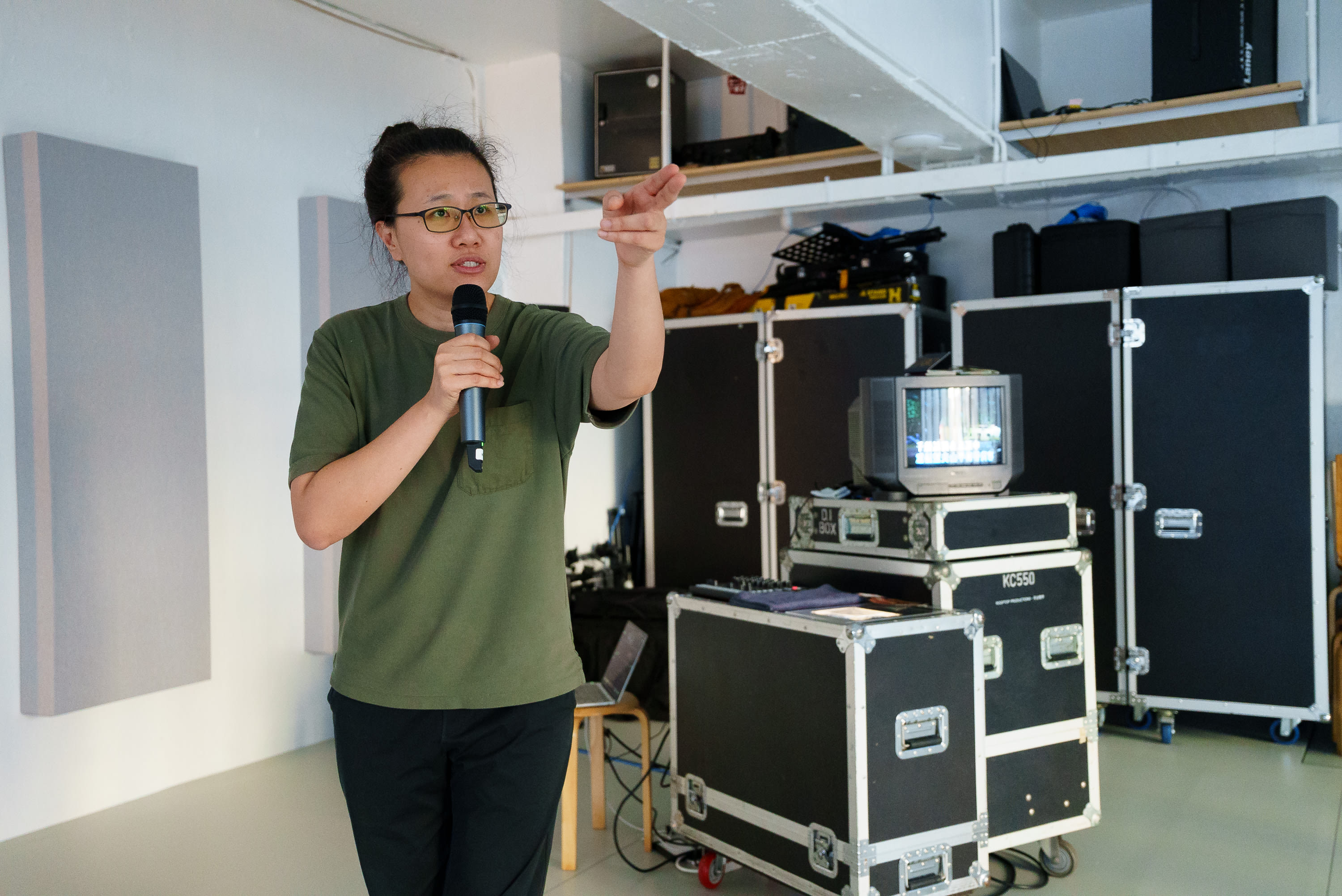 演員李婉晶在天台製作的當代劇場作品《卡啦OK》排練現場，手持麥克風並指向前方，身後擺放著演出用的飛行箱和器材。《卡啦OK》排練。