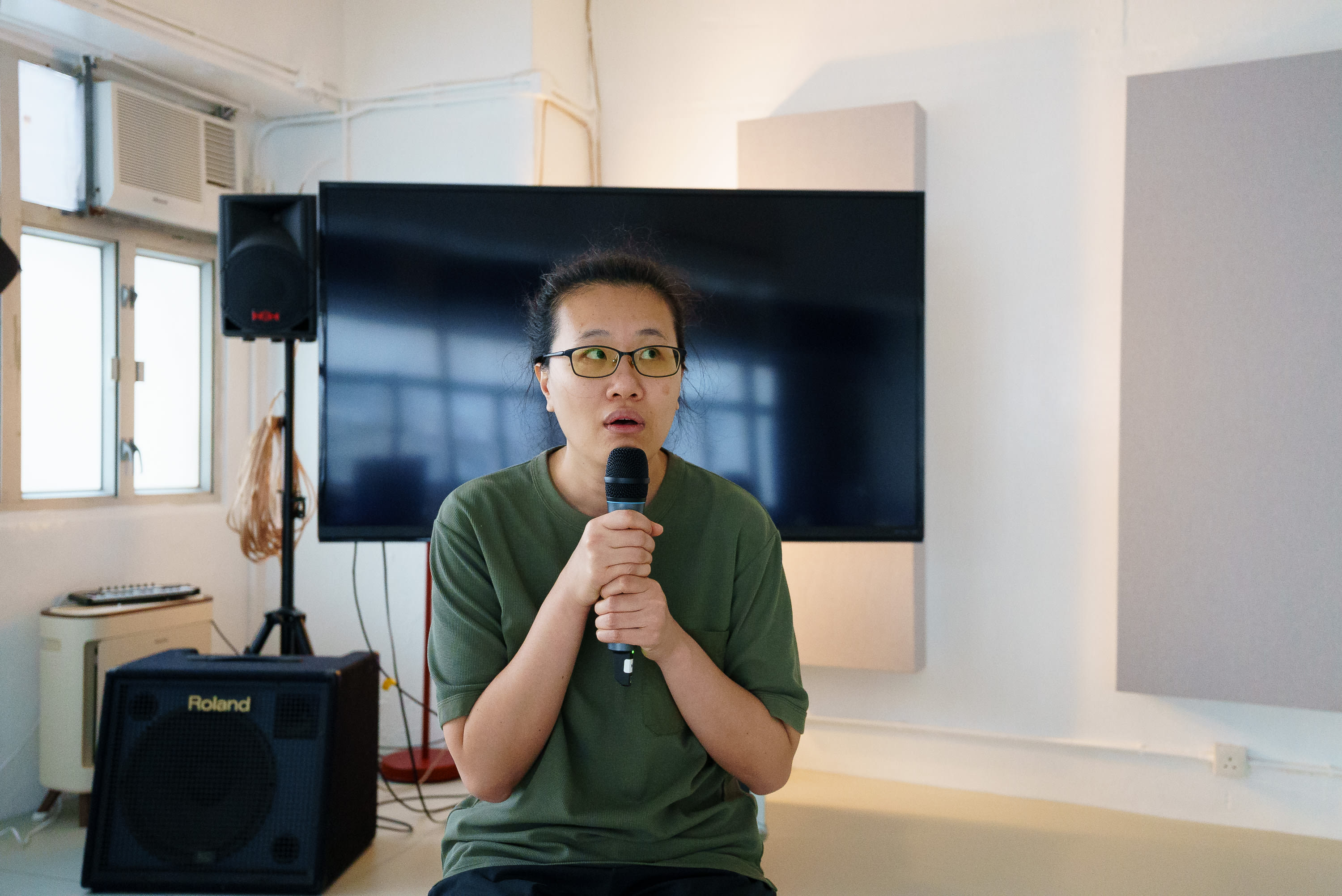 Michelle Li holds a microphone during a rehearsal for Rooftop Productions' contemporary Hong Kong theatre piece, 'Karaoke' (《卡啦OK》). She sits in a studio space with professional stage equipment, including a Roland amplifier and sound monitor, capturing an intimate, focused moment of artistic preparation. 'Karaoke rehearsals'