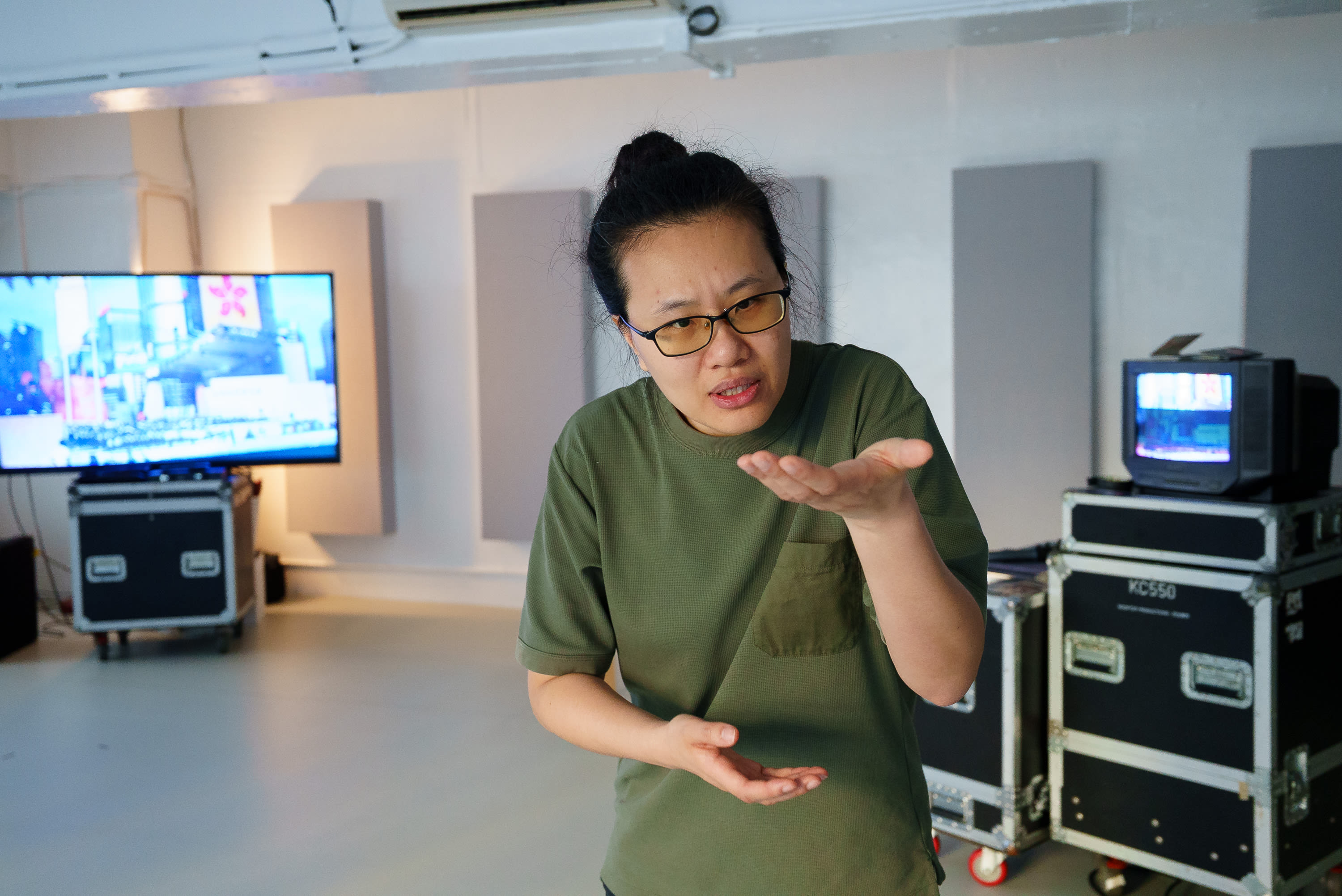 李婉晶站在排練室中，正在進行天台製作（Rooftop Productions）當代劇場作品《卡啦OK》的排練。她佩戴著琥珀色眼鏡，身穿橄欖綠色T恤，雙手向上攤開，呈現出彷彿在講述故事或展現角色內心掙扎的動態。背景中可以看到專業的錄音室隔音板、播放影像的大型螢幕以及舞台製作專用的器材箱，營造出沉浸式劇場表演的氛围。《卡啦OK》排練。