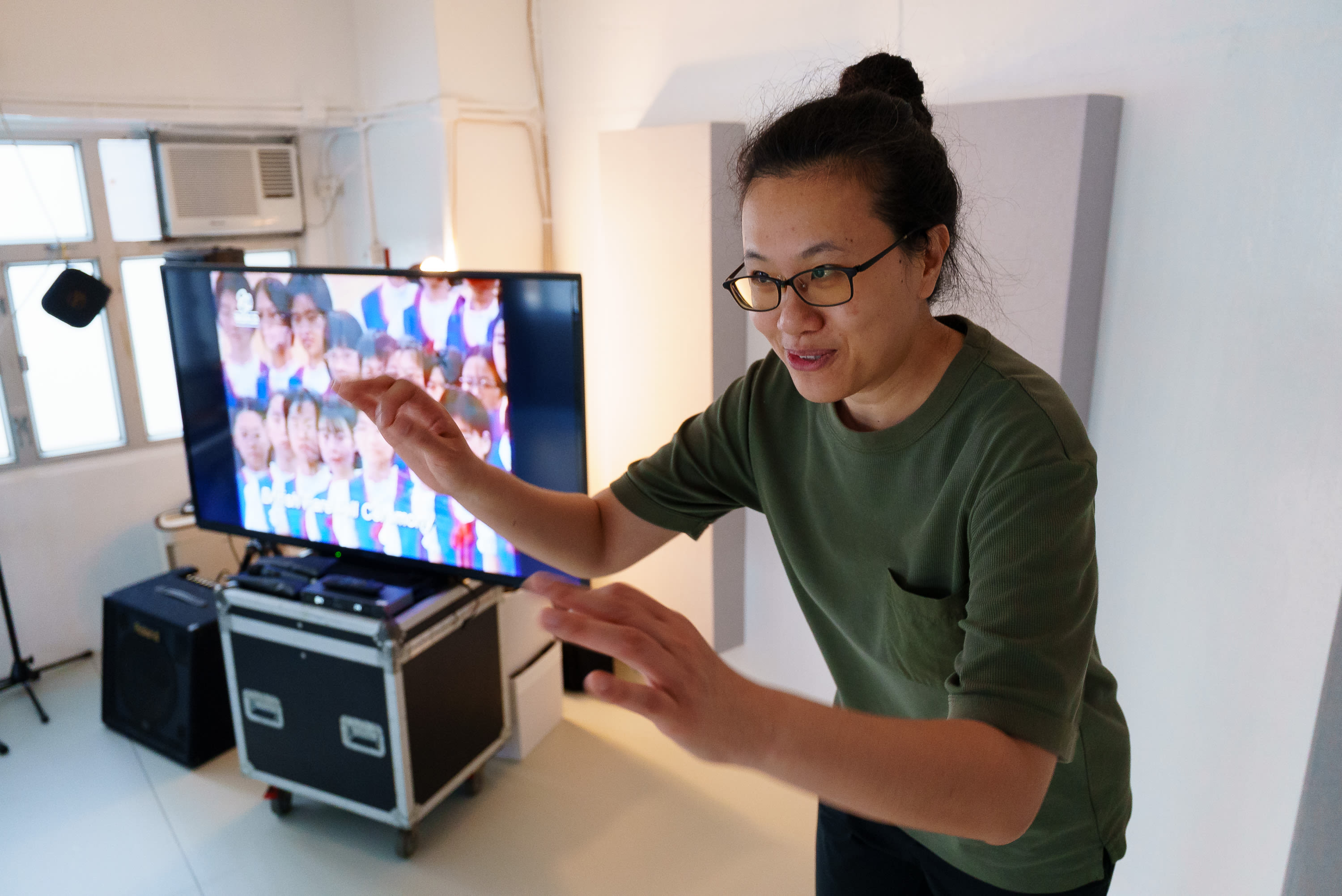In a rehearsal space for Rooftop Productions' contemporary theatre piece 'Karaoke', performer Michelle Li stands in front of a flat-screen television displaying a grid of choir members. She wears glasses and an olive green t-shirt, her hands outstretched as if conducting or guiding the performance with focused intensity. The setting captures a moment of creative development in the Hong Kong independent arts scene. 'Karaoke rehearsals'